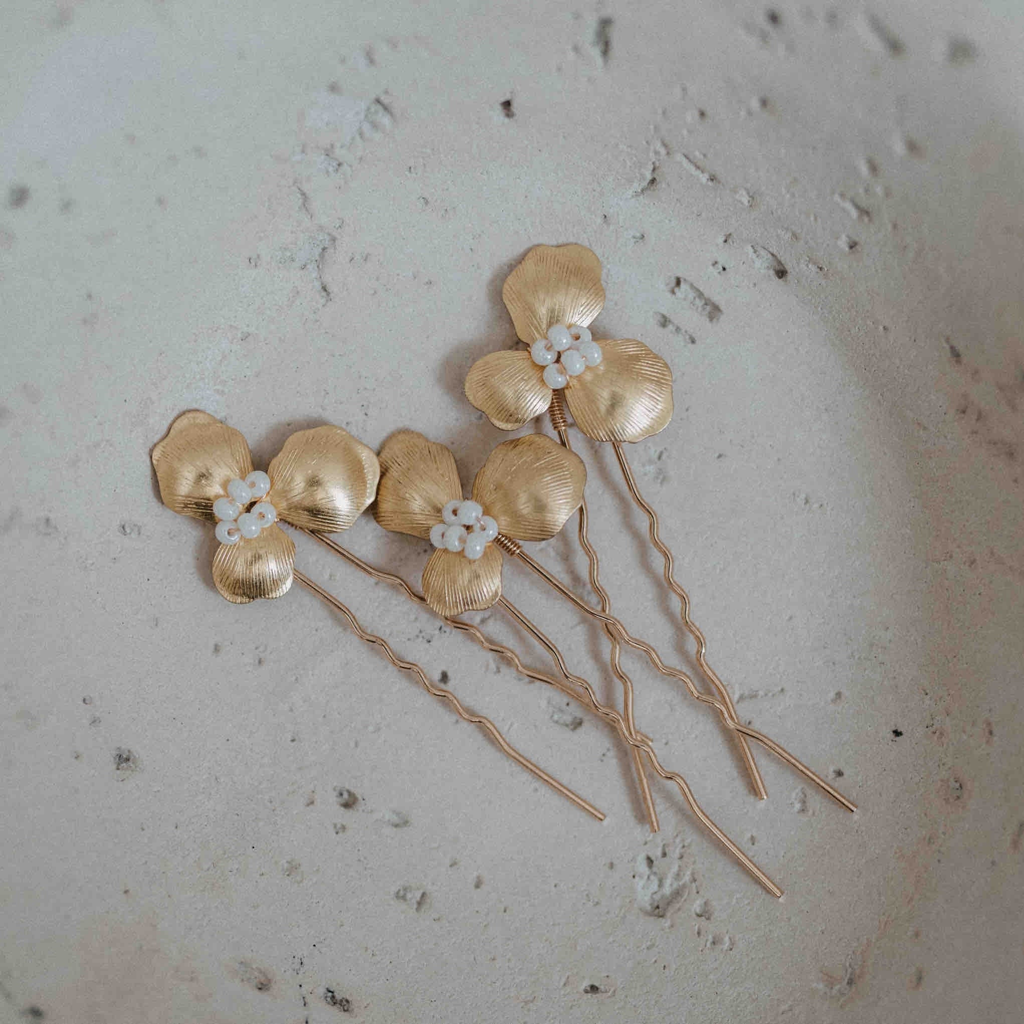 Drei Haarnadeln mit goldenen Blüten von Fräulein Maya, verziert mit strukturierten Metallblüten und weißen Blumen-Details, sind diagonal auf einer rauen, cremefarbenen Oberfläche angeordnet und ergänzen stilvoll deine Haarschmuckkollektion.