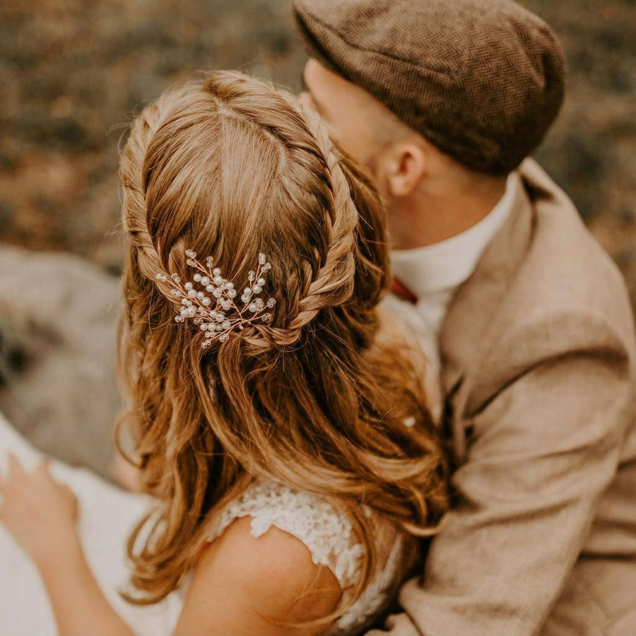 Eine Frau mit gewelltem, hellbraunem Haar, das zu einem Zopf mit einem Haarschmuck aus Perlen und Kristallen gestylt ist, sitzt neben einem Mann, der eine braune Schiebermütze und einen beigen Anzug trägt. Sie stehen dicht beieinander, von der Kamera abgewandt, in einer Außenansicht.