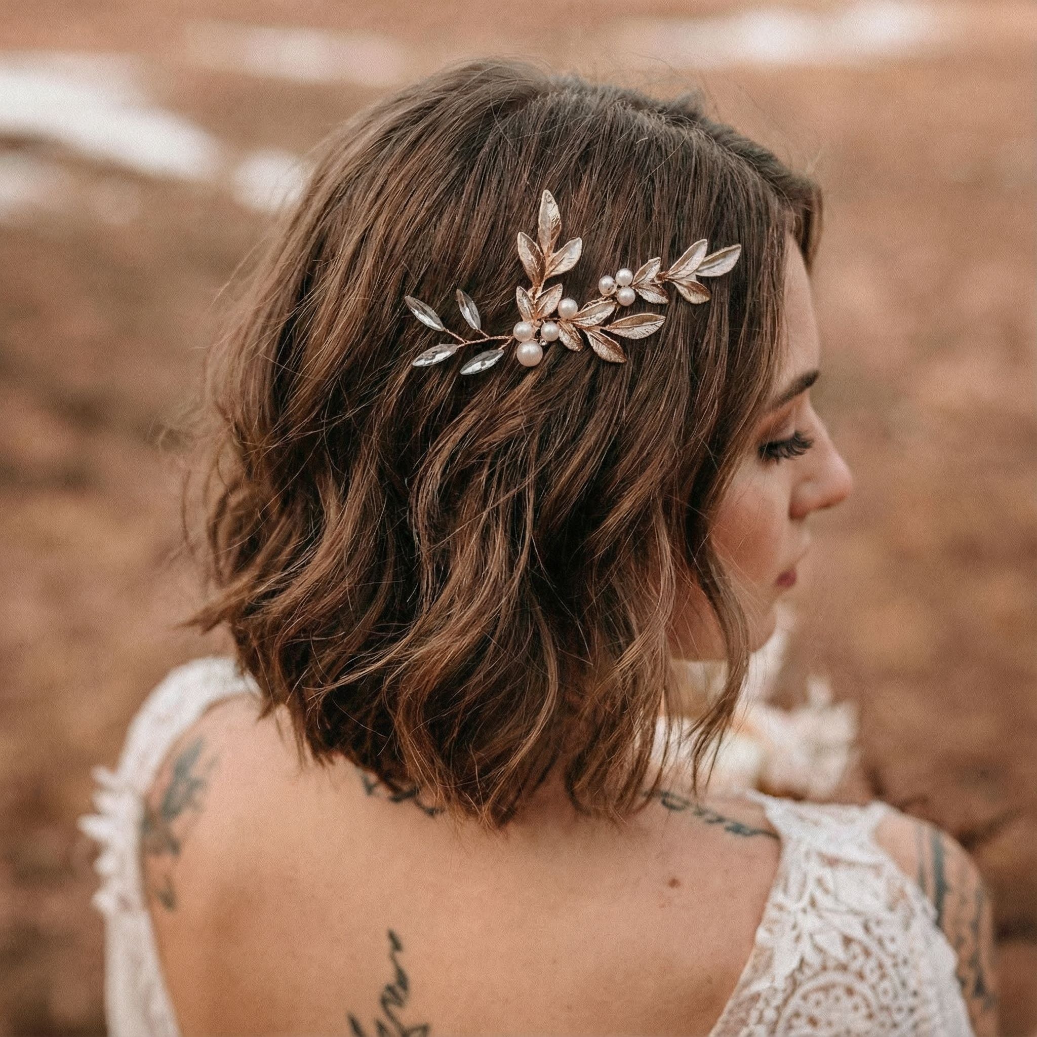 Eine Frau mit kurzen, gewellten braunen Haaren trägt einen Haarschmuck aus Gold und Perlenblättern. Sie trägt ein weißes Spitzenoberteil mit floralen Tattoos auf der Schulter. Der Hintergrund ist in sanften Braun- und Beigetönen gehalten, die eine Umgebung im Freien andeuten.
