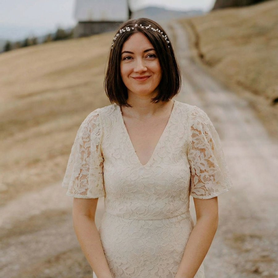 Eine Frau mit einem kurzen braunen Bob und einem Perlenstirnband steht draußen auf einem Feldweg und lächelt. Sie trägt ein cremefarbenes Spitzenkleid mit Flatterärmeln. Im Hintergrund sind braune grasbewachsene Hügel, verschwommene Bäume und ein entferntes weißes Gebäude zu sehen.
