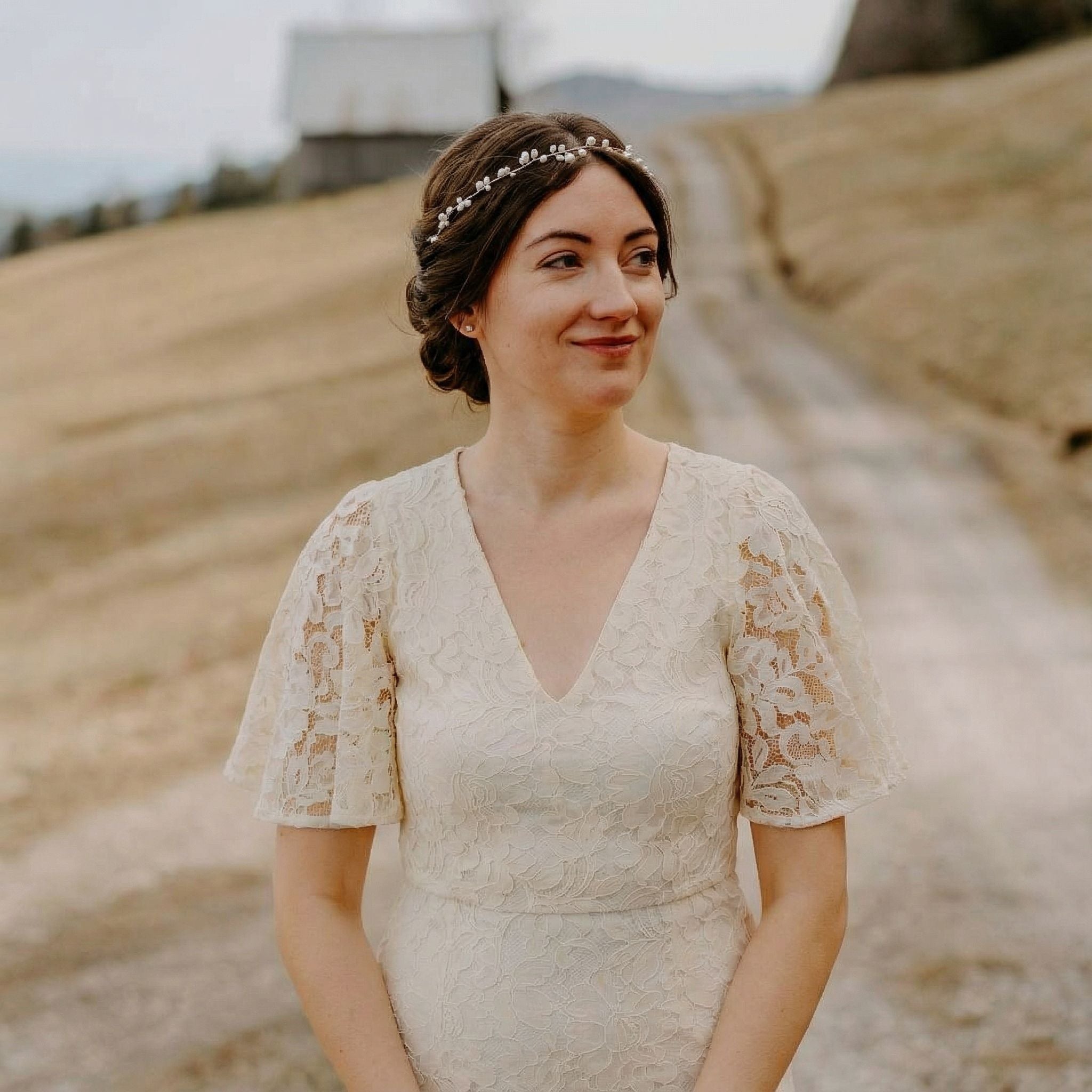 Eine Frau in einem spitzenbesetzten, kurzärmeligen weißen Kleid steht auf einem Feldweg im Freien, mit sanften Hügeln und einem rustikalen Holzgebäude im Hintergrund. Sie hat braunes Haar, das mit einem zarten Haarreif zurückgestylt ist, lächelt sanft und blickt zur Seite.