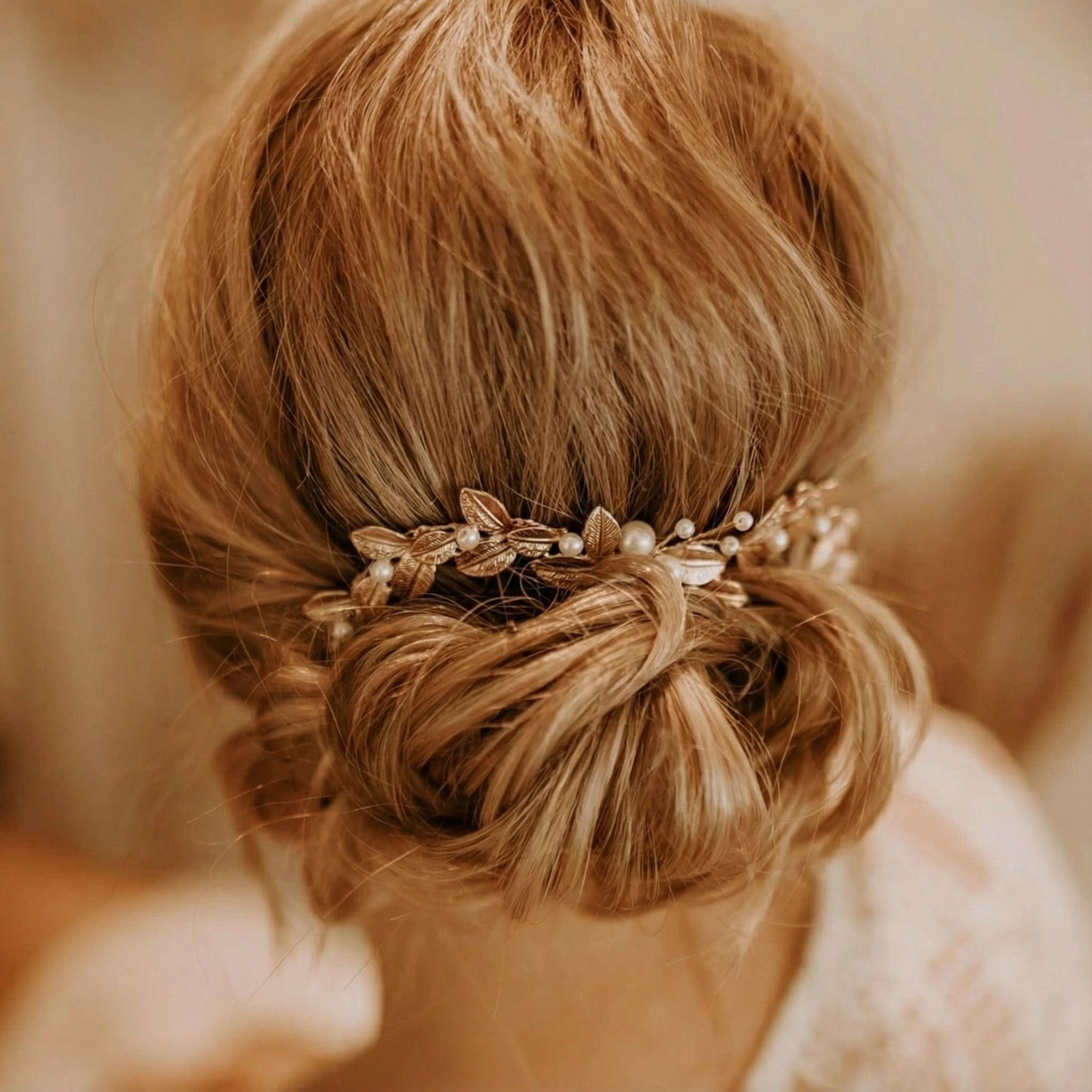 Eine Frau mit blondem Haar, das zu einem niedrigen, lockeren Dutt gestylt ist, geschmückt mit einem komplizierten Haarschmuck aus Gold und Perlen, der Blätter und kleine Perlen enthält. Der Hintergrund ist sanft verschwommen und lenkt die Aufmerksamkeit auf die elegante Frisur.