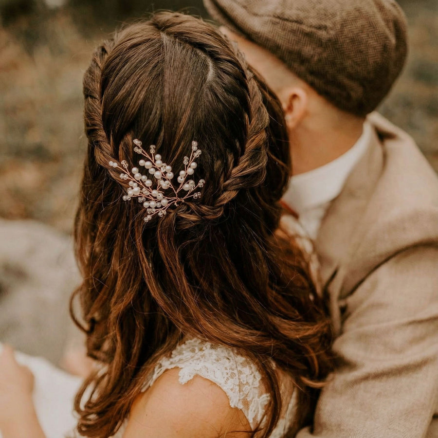 Eine Frau mit langen braunen Haaren, die in lockeren Wellen und zu einem Zopf geflochten sind und mit einem zarten Haarschmuck aus kleinen weißen Perlen geschmückt sind, sitzt dicht neben einem Mann mit brauner Schiebermütze und hellbraunem Anzug. Beide schauen von der Kamera weg und befinden sich im Freien.