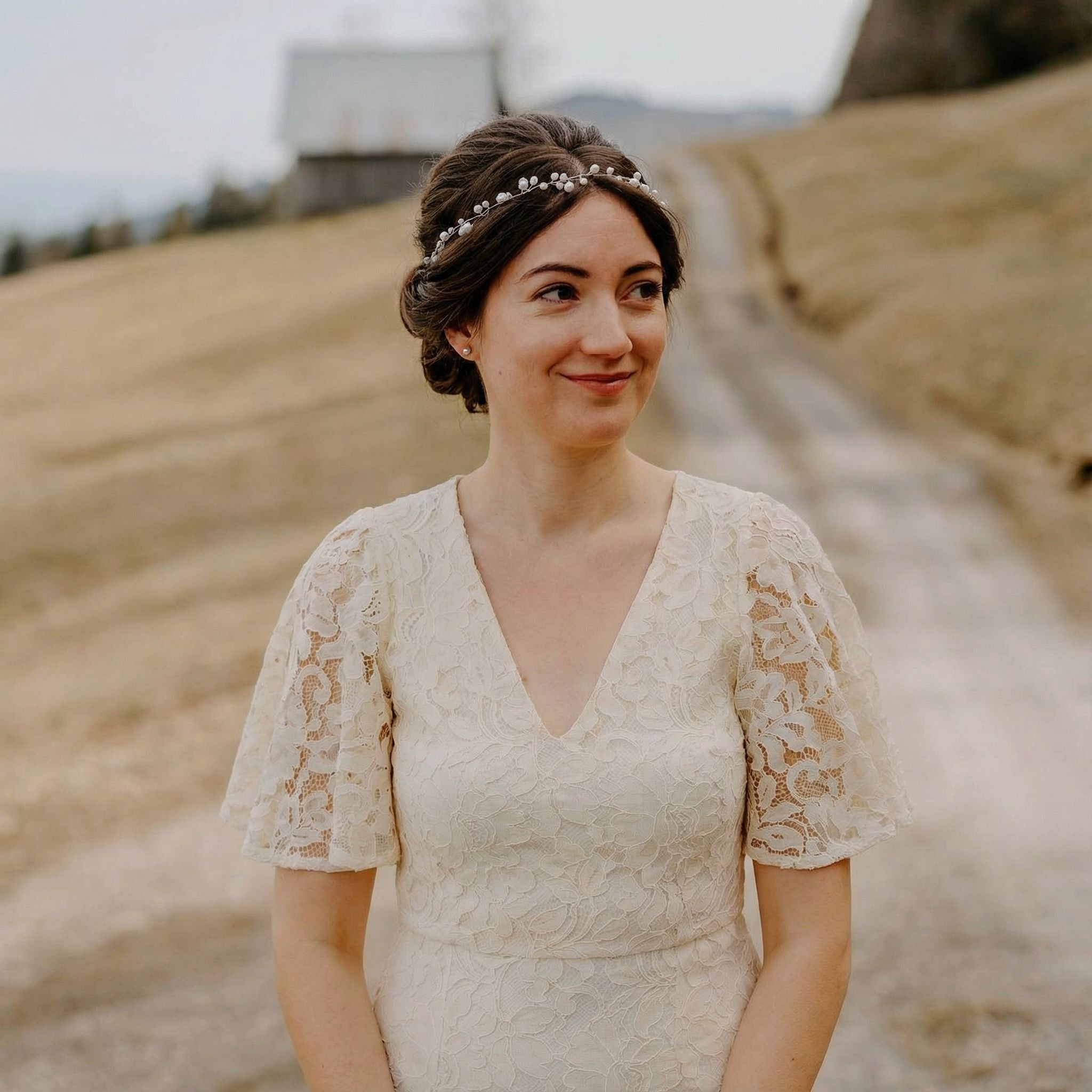 Eine Frau in einem weißen Spitzenkleid mit Flatterärmeln steht auf einem Feldweg. Sie hat helle Haut, braunes Haar, das mit einem Perlenstirnband frisiert ist, und schaut leicht lächelnd zur Seite. Der Hintergrund ist eine ländliche Landschaft mit einer Scheune und grasbewachsenen Hügeln.