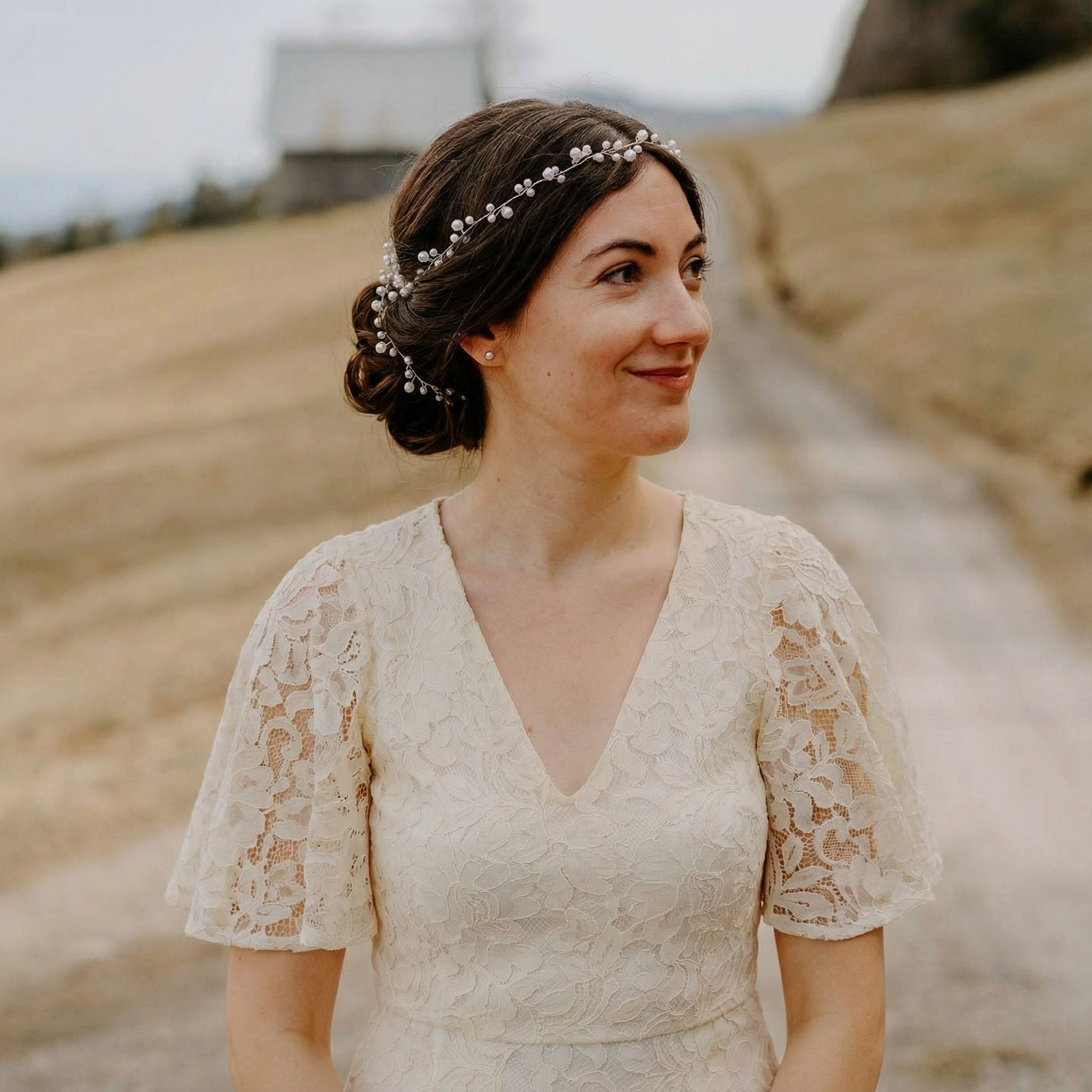 Eine Frau in einem cremefarbenen Spitzenkleid steht im Freien auf einem Feldweg, lächelt und blickt zur Seite. Ihr Haar ist zu einem tiefen Dutt mit einem zarten Perlenstirnband frisiert. Der Hintergrund zeigt ein grasbewachsenes Feld, eine Scheune und Hügel unter einem bewölkten Himmel.