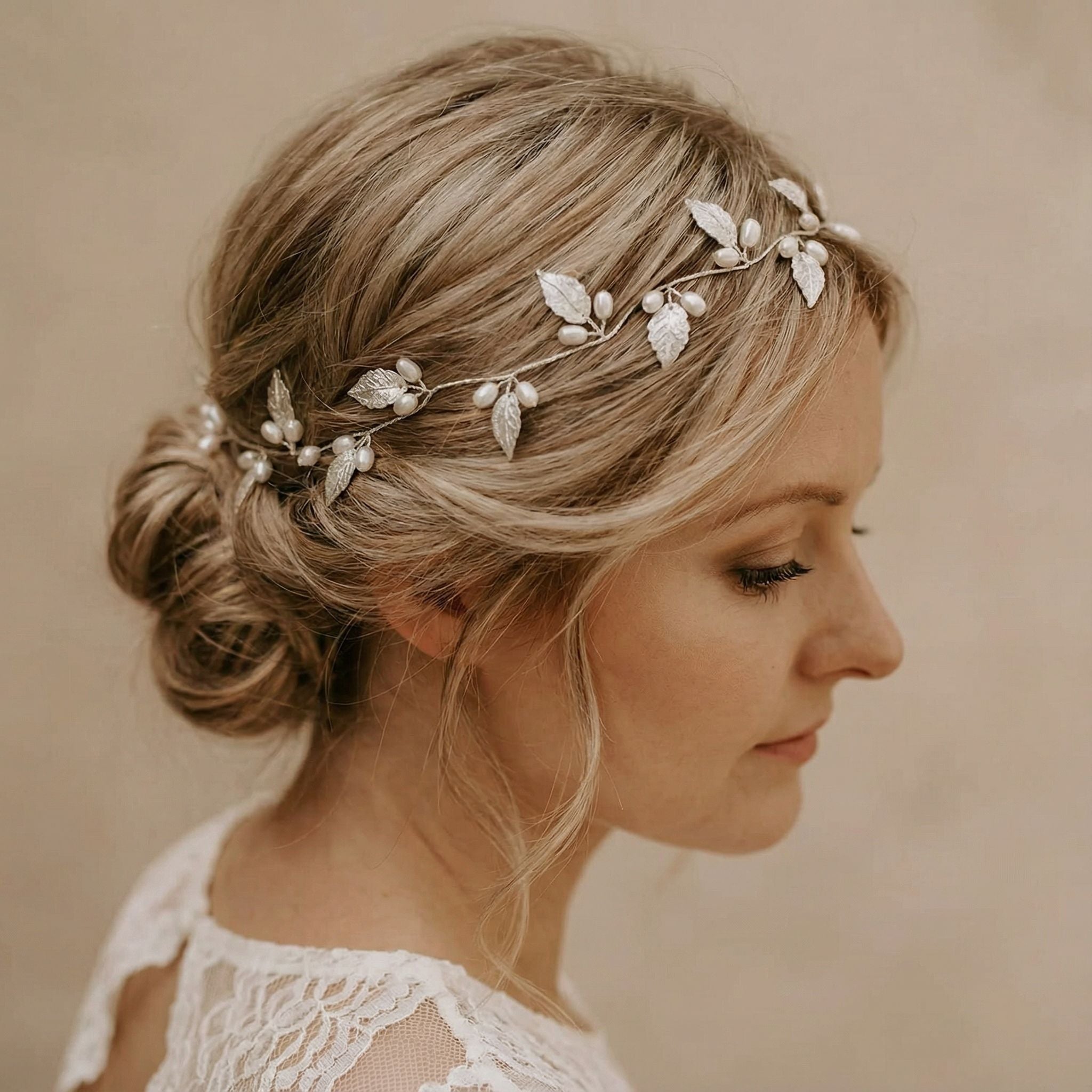 Eine Frau mit blondem Haar, das zu einem lockeren, niedrigen Dutt frisiert ist, trägt einen zarten silbernen Kopfschmuck mit Blatt- und Perlendetails. Sie ist im Profil, nach rechts gewandt, und trägt ein Spitzenkleid. Der Hintergrund ist in zartem Beige gehalten, was einen sanften, eleganten Eindruck vermittelt.