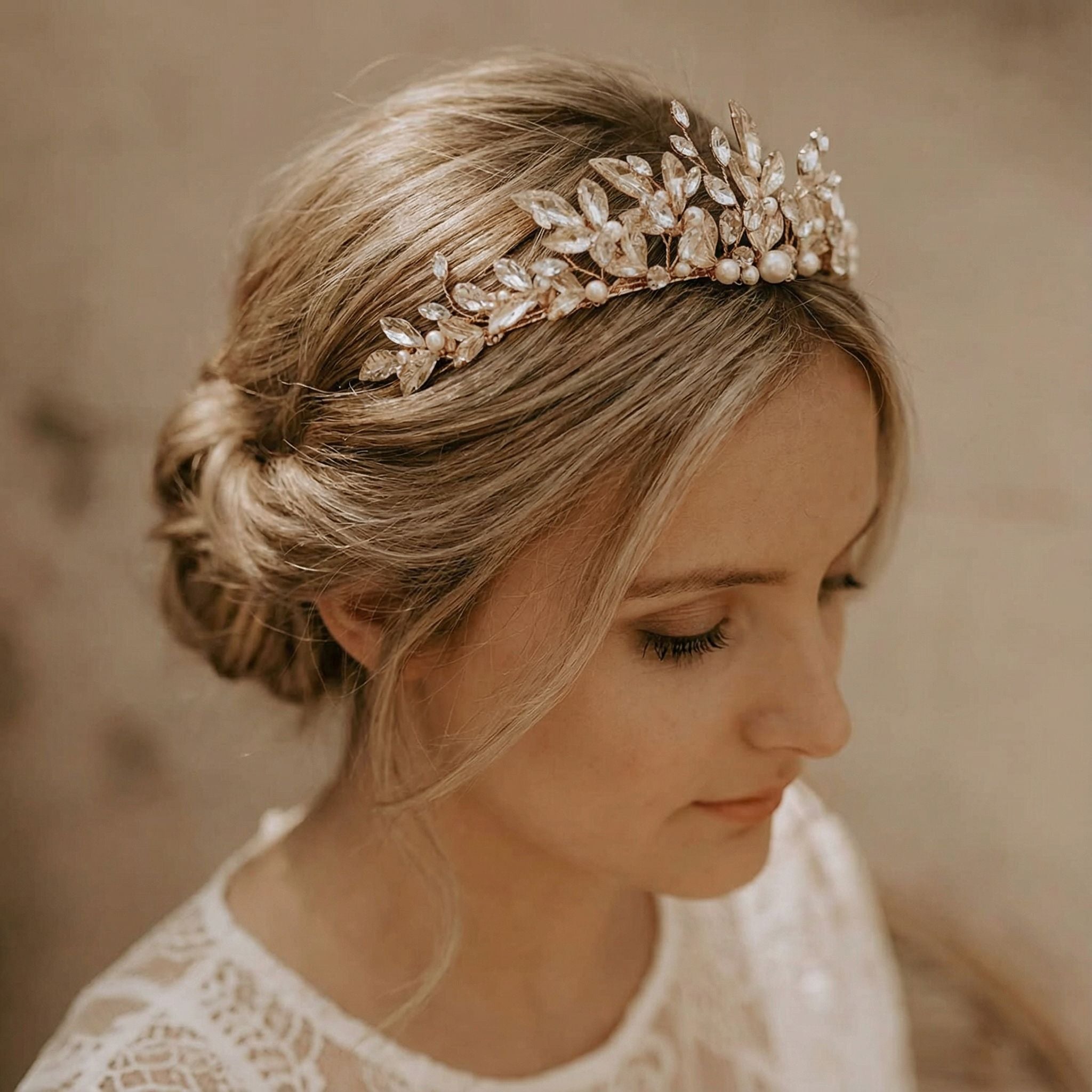 Eine Frau mit blondem Haar, das zu einem niedrigen, lockeren Dutt frisiert ist, trägt ein zartes goldenes Diadem, das mit Perlen und klaren Kristallen verziert ist. Sie trägt ein weißes Spitzengewand und blickt sanft nach unten, was ihr einen ruhigen und eleganten Brautlook verleiht.