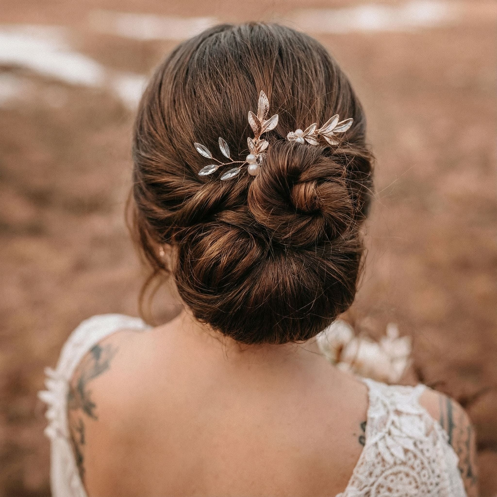 Eine Frau mit braunem Haar, das zu einem eleganten Dutt frisiert und mit goldenen und perlenbesetzten Haarnadeln verziert ist. Sie trägt ein weißes Spitzenkleid mit floralen Mustern und hat Tätowierungen auf den Schultern. Der Hintergrund ist unscharf und zeigt erdige Farbtöne im Freien.