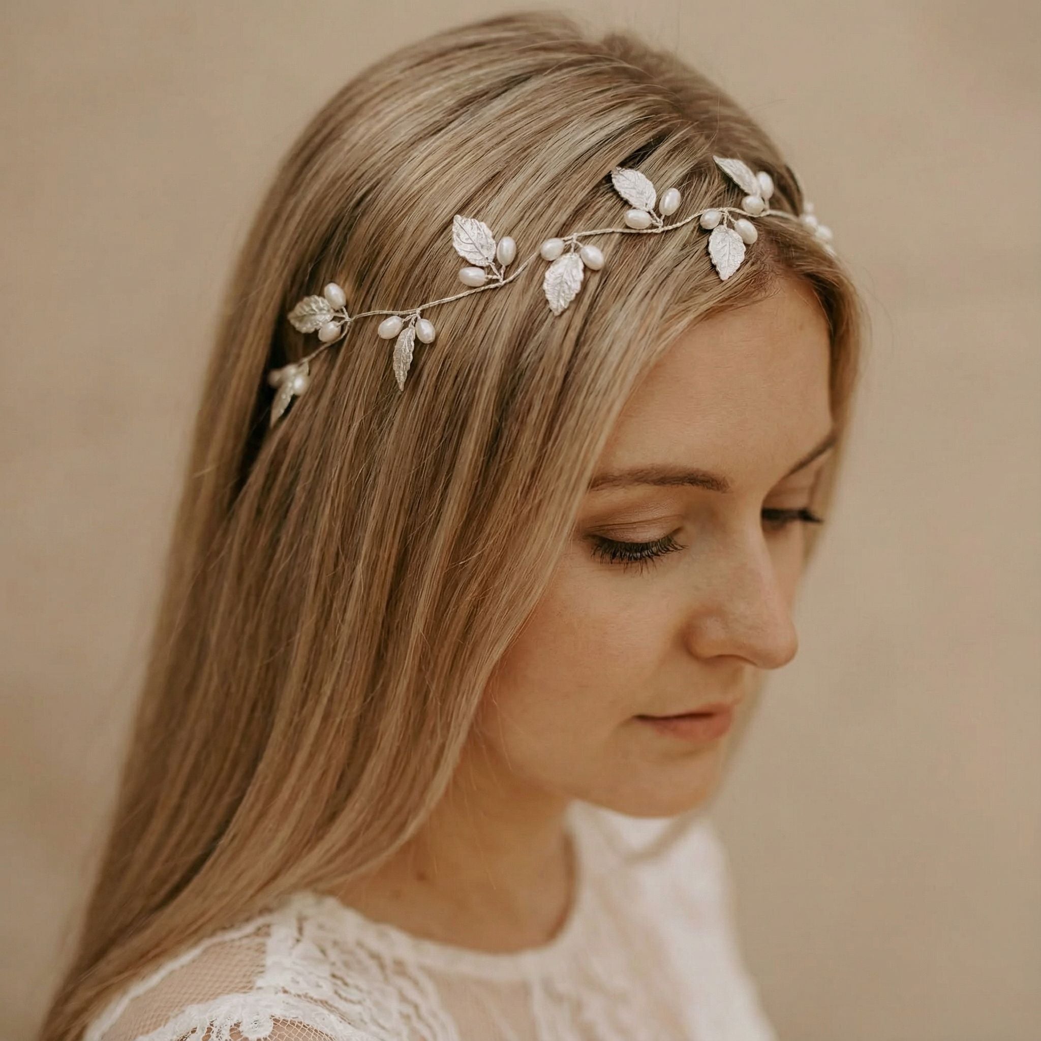 Eine Frau mit langem, glattem blondem Haar trägt einen zarten Kopfschmuck, der mit silbernen Blättern und kleinen Perlen verziert ist. Sie ist mit einem weißen Spitzenkleid bekleidet und blickt vor einem neutralen Hintergrund mit heiterer Miene nach unten.