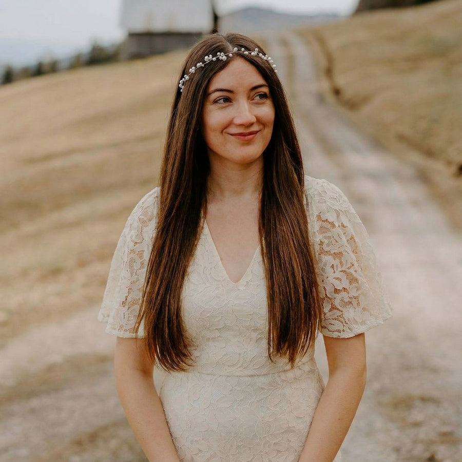Eine Frau mit langem, glattem, braunem Haar, die ein zartes, perlenbesetztes Stirnband und ein weißes Spitzenkleid mit Flatterärmeln trägt, steht im Freien auf einem Feldweg mit trockenem Gras auf beiden Seiten. Sie lächelt sanft und blickt leicht nach links.