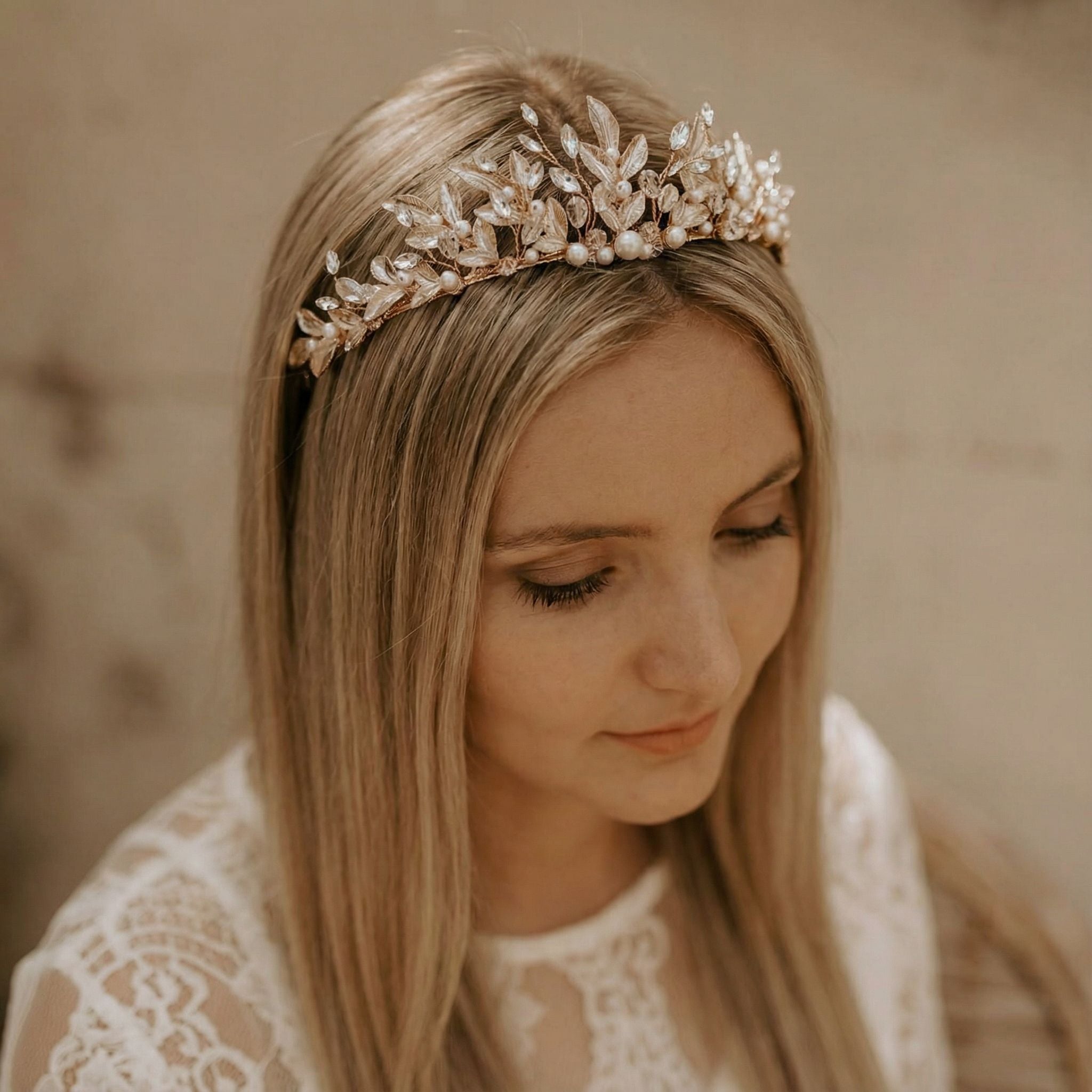 Eine Frau mit glattem, blondem Haar trägt ein zartes, mit Gold und Perlen besetztes Blumenstirnband. Sie blickt mit heiterer Miene nach unten. Sie trägt ein weißes Spitzengewand vor einem sanft beleuchteten, neutralen Hintergrund.