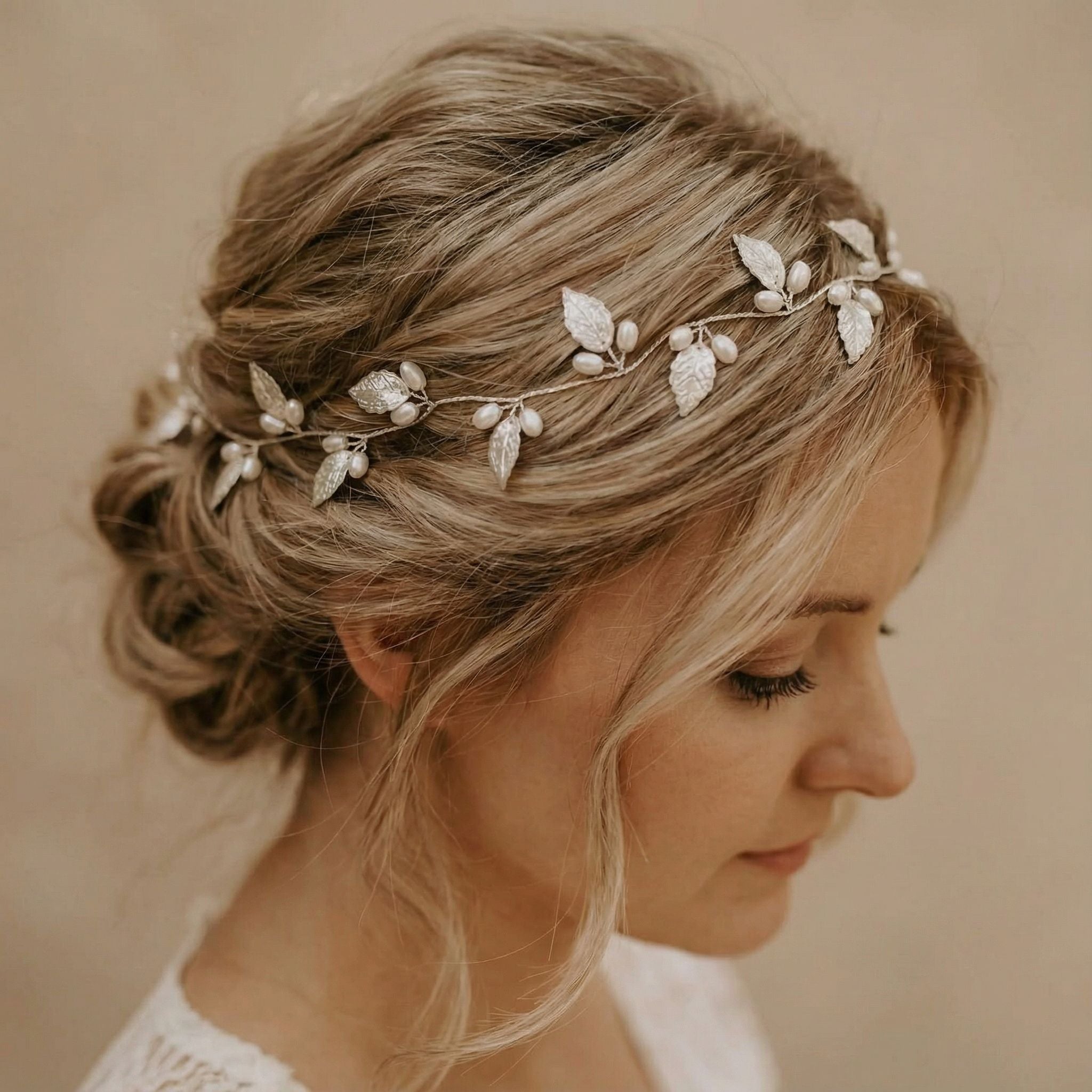 Eine Frau mit blondem Haar, das zu einer lockeren, eleganten Hochsteckfrisur frisiert und mit einem zarten Kopfschmuck aus silbernen Blättern und kleinen Perlen geschmückt ist. Sie ist im Profil vor einem neutralen Hintergrund dargestellt, trägt ein leichtes Spitzengewand und blickt sanft nach unten.