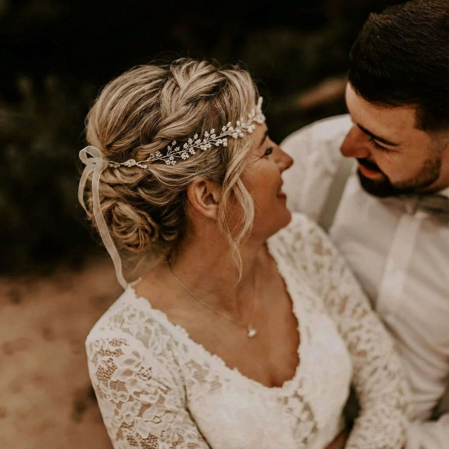Eine Braut mit geflochtenen blonden Haaren, die mit einem zarten Blumenkopfschmuck und einem Spitzenkleid geschmückt sind, lächelt einen bärtigen Bräutigam mit weißem Hemd und Fliege herzlich an. Sie stehen dicht beieinander und teilen einen intimen, freudigen Moment im Freien.