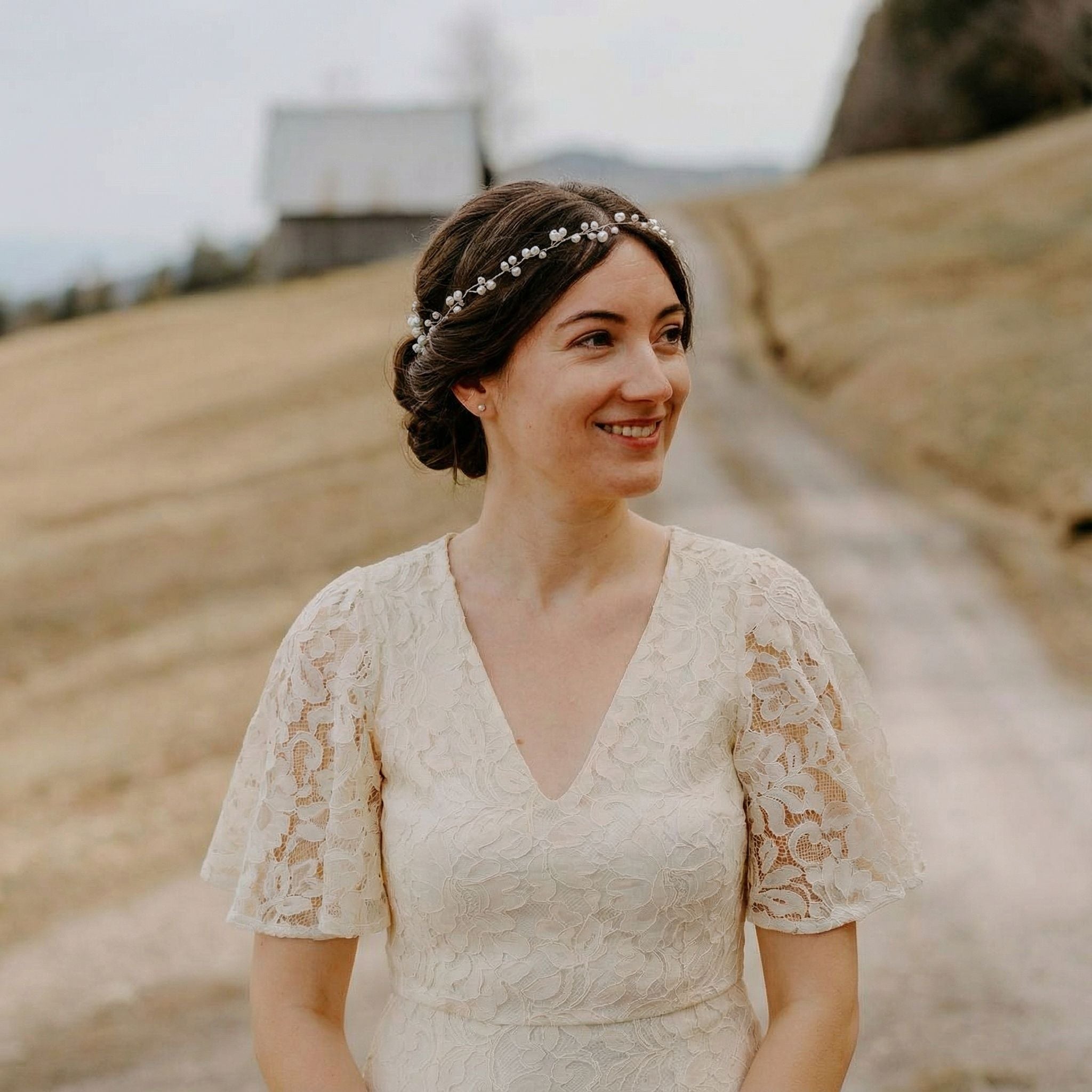 Eine Frau in einem elfenbeinfarbenen Spitzenkleid mit Flatterärmeln steht auf einem Feldweg im Freien. Sie lächelt und blickt zur Seite. Ihr dunkles Haar ist zu einem tiefen Dutt mit einem Perlenstirnband frisiert. Der Hintergrund ist eine ländliche Landschaft mit einem grasbewachsenen Hügel und einer rustikalen Scheune.