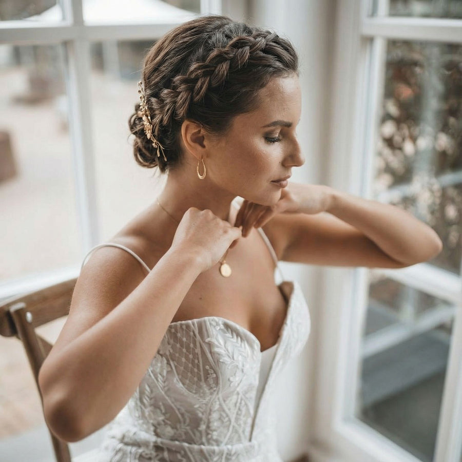 Eine Frau in einem weißen, strukturierten, ärmellosen Hochzeitskleid sitzt an einem Fenster. Sie hat ihr braunes Haar zu einer geflochtenen Hochsteckfrisur mit einem goldenen Haarteil frisiert und trägt Reifohrringe und eine Anhänger-Halskette, die sie zurechtrückt, während sie gelassen nach unten schaut.