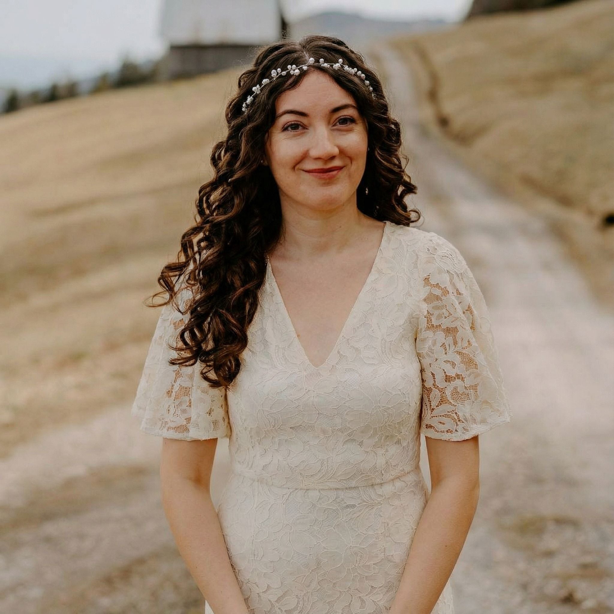 Eine Frau mit langen, lockigen braunen Haaren trägt ein zartes Blumenstirnband und ein cremefarbenes Spitzenkleid mit Flatterärmeln. Sie steht im Freien auf einem Feldweg mit beigen Grashügeln und einem unscharfen Gebäude im Hintergrund und lächelt sanft in die Kamera.
