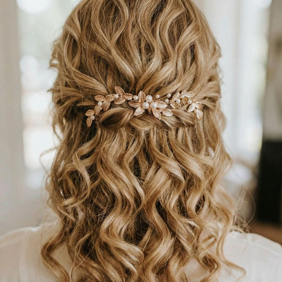 Eine Frau mit langem, blondem, gewelltem Haar, das halb hochgesteckt und halb heruntergelassen ist. Ihr Haar ist mit einem verschnörkelten Haarteil mit goldenen Blättern und weißen Perlen geschmückt, das ihrer sanft gelockten Frisur einen eleganten Touch verleiht. Der Hintergrund ist sanft verschwommen.