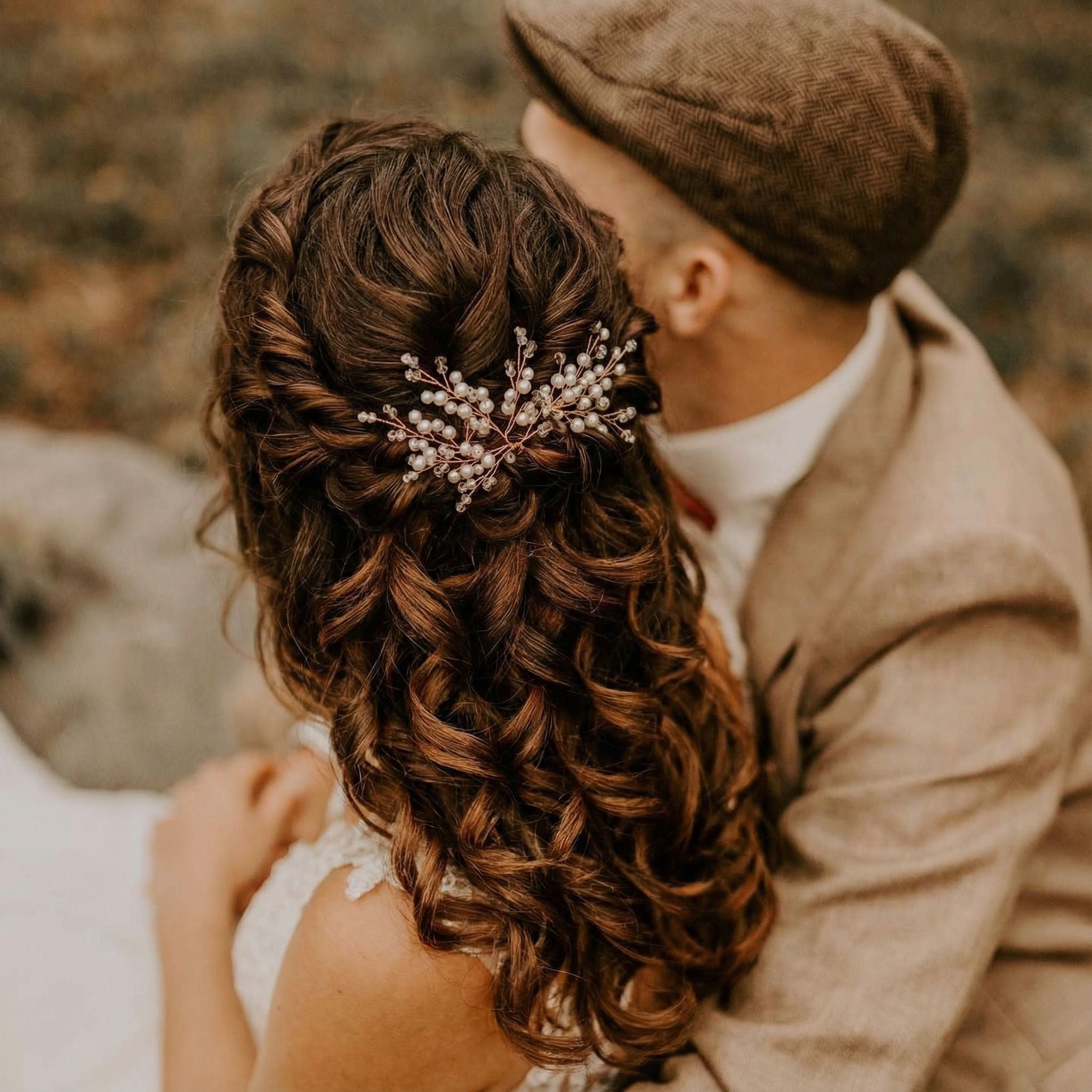 Ein Paar sitzt eng beieinander im Freien. Die Frau in einem weißen Kleid trägt langes, braunes, lockiges Haar, das mit einem Haarteil aus Perlen und Kristallen halb hochgesteckt ist. Der Mann, der eine braune Schiebermütze und einen beigen Anzug trägt, hat seinen Arm um sie gelegt und schaut von der Kamera weg.