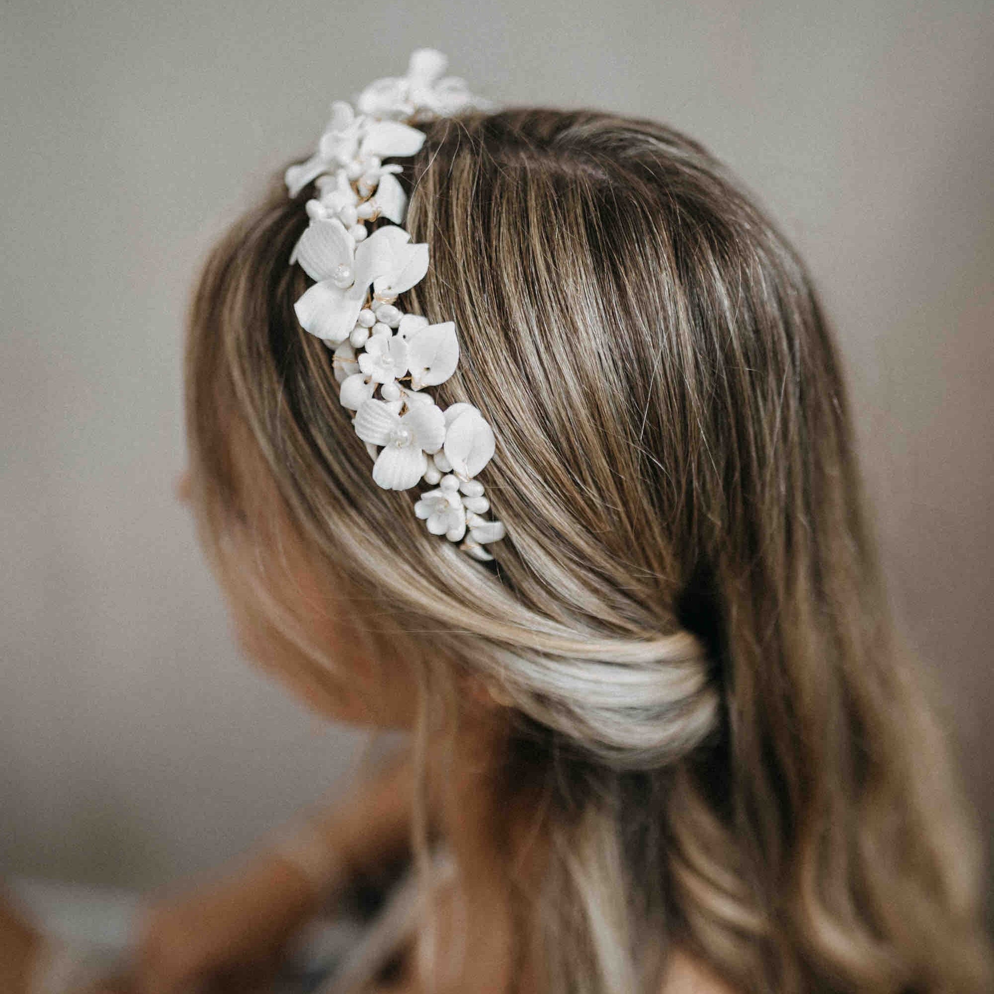 Eine Frau mit gewelltem, hellbraunem Haar trägt das Stirnband Fräulein Maya Krone Braut mit Keramik Blüten, gestylt zu einer eleganten Hochsteckfrisur für die Braut. Ihr schulterfreies Spitzenkleid ist teilweise vor einem sanft verschwommenen Hintergrund zu sehen.