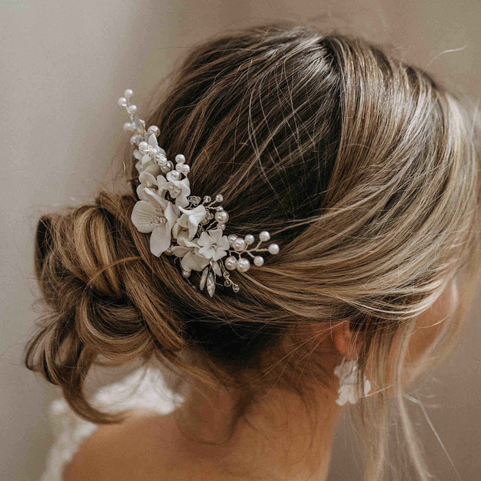 Eine Frau mit blondem Haar in einem lockeren, niedrigen Dutt, geschmückt mit dem Fräulein Maya Haarkamm mit Keramik Blumen und Kristallen. Sie trägt ein leichtes Spitzenkleid und Perlenohrringe, von hinten fotografiert.