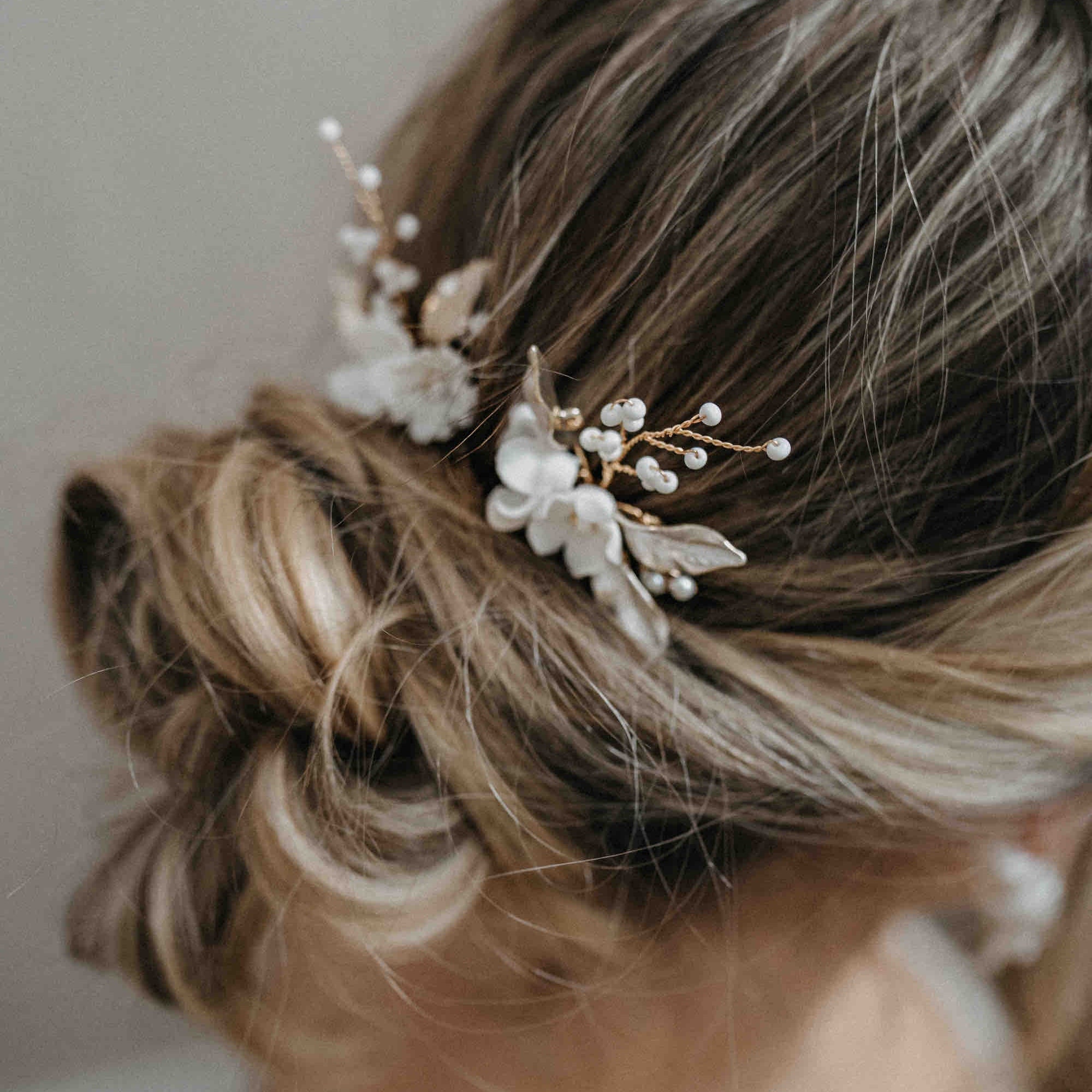 Eine Nahaufnahme der eleganten Brautfrisur einer Frau, die zu einem lockeren Dutt gestylt und mit Fräulein Maya Haarnadeln mit Keramik Blumen und Blättern (3 Stück) geschmückt ist. Die zarten weißen Blüten und goldenen Blätter heben sich von dem sanft verschwommenen Hintergrund ab.