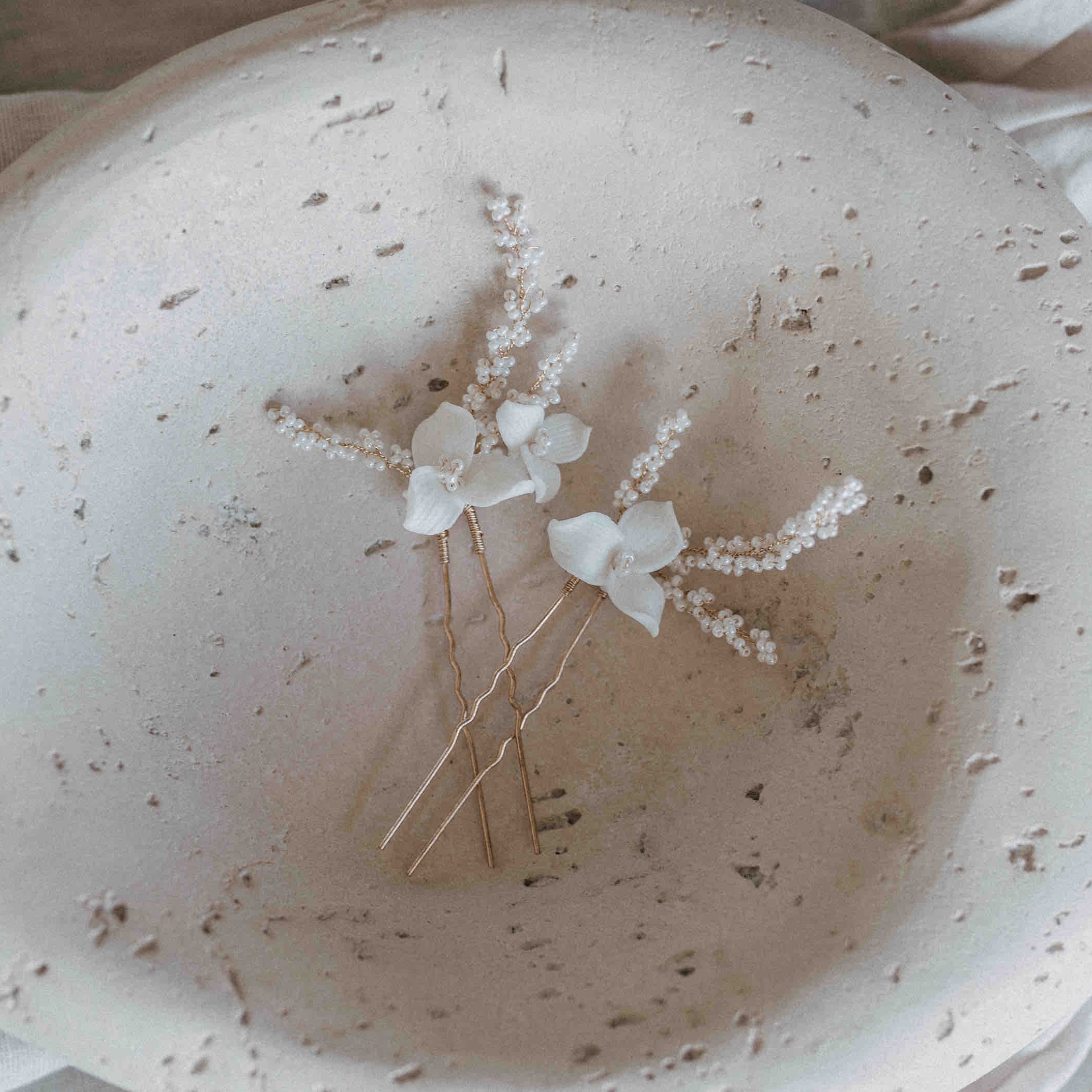 Zwei goldene Fräulein Maya Keramik Haarnadeln mit Blumen und feinen Perlen (2 Stück) liegen in einer strukturierten, cremefarbenen Schale mit kleinen Löchern und einem handgefertigten Look. Weicher Stoff lugt im Hintergrund hervor.