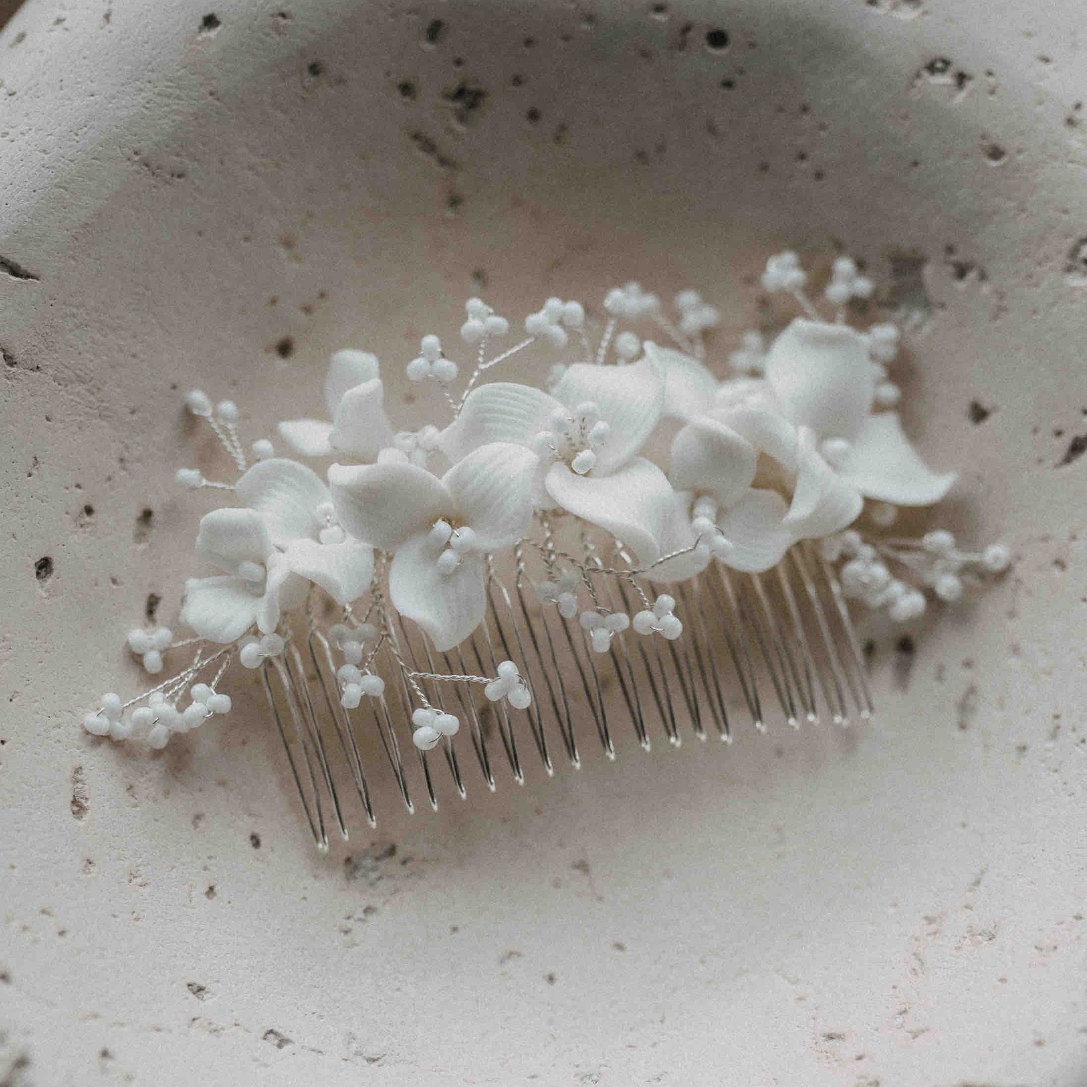 Der Keramik Haarkamm mit Blumen und Perlen von Fräulein Maya zeigt weiße Kunstblumen und kleine Perlen auf cremefarbenem Stein. Mit sichtbaren Metallzähnen - das perfekte Haar-Accessoire für deine Brautfrisur.