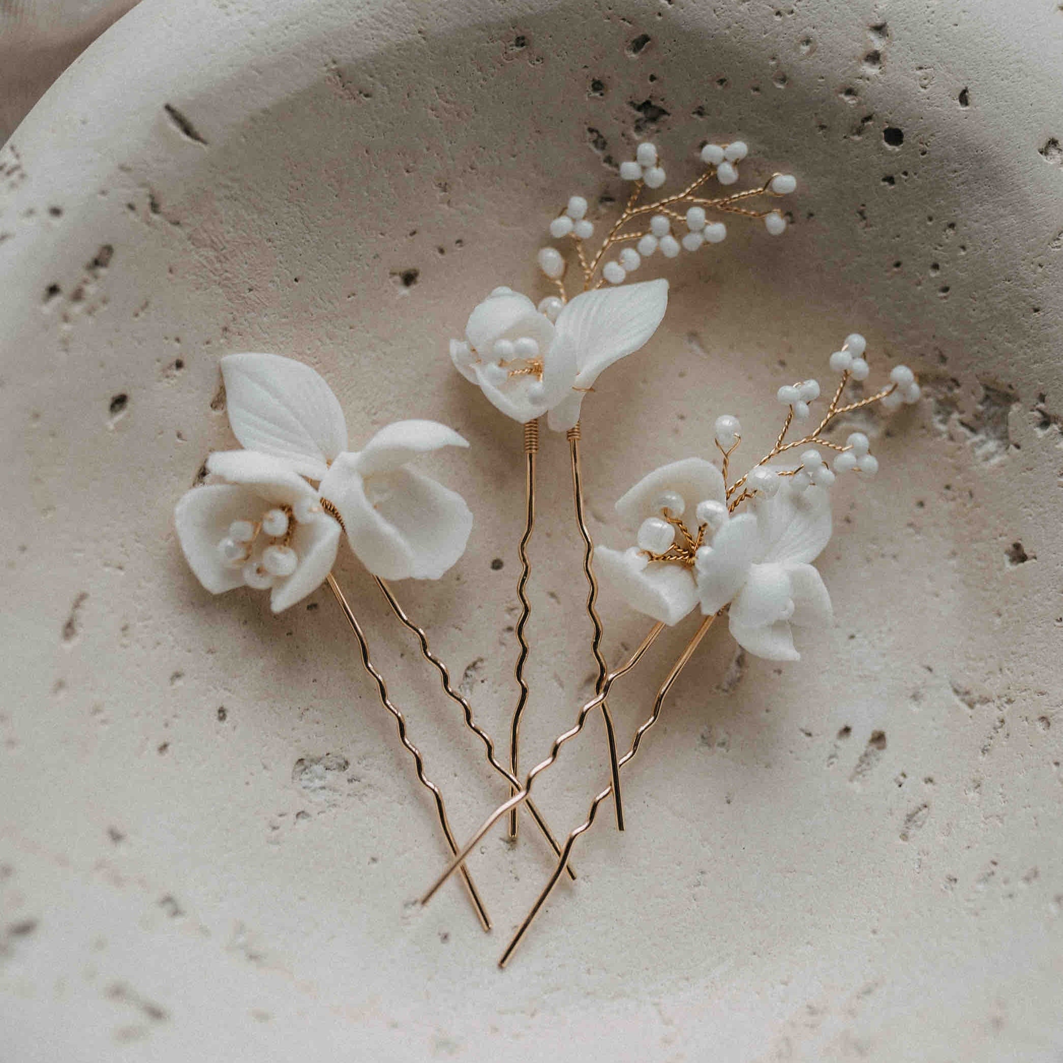 Drei Haarnadeln mit großen Keramik Blumen (3 Stück) von Fräulein Maya, verziert mit goldenen Stielen und zarten, perlenähnlichen Details, liegen auf einer strukturierten Schale - stilvolle botanische Haaraccessoires für die Braut.