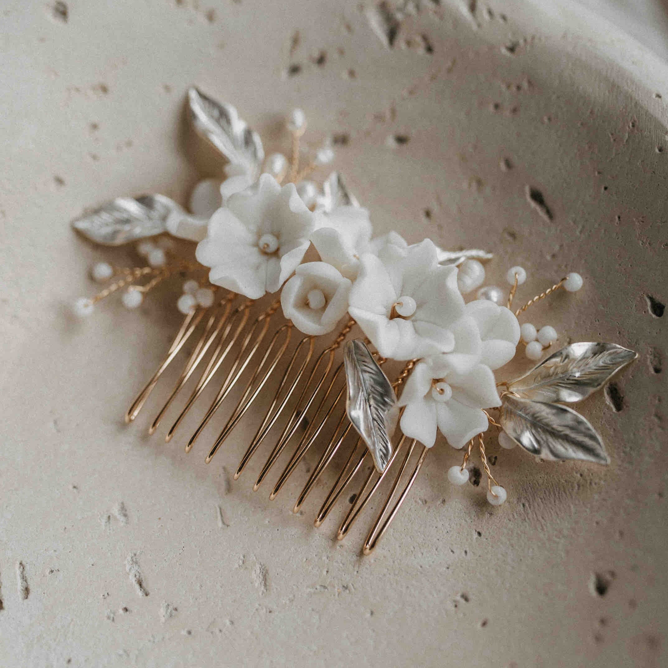 Der Haarkamm mit Keramik Blumen und Blätter von Fräulein Maya, verziert mit weißen Keramikblüten, perlenartigen Details und silbernen Blättern, präsentiert sich elegant auf einer strukturierten beigefarbenen Oberfläche.