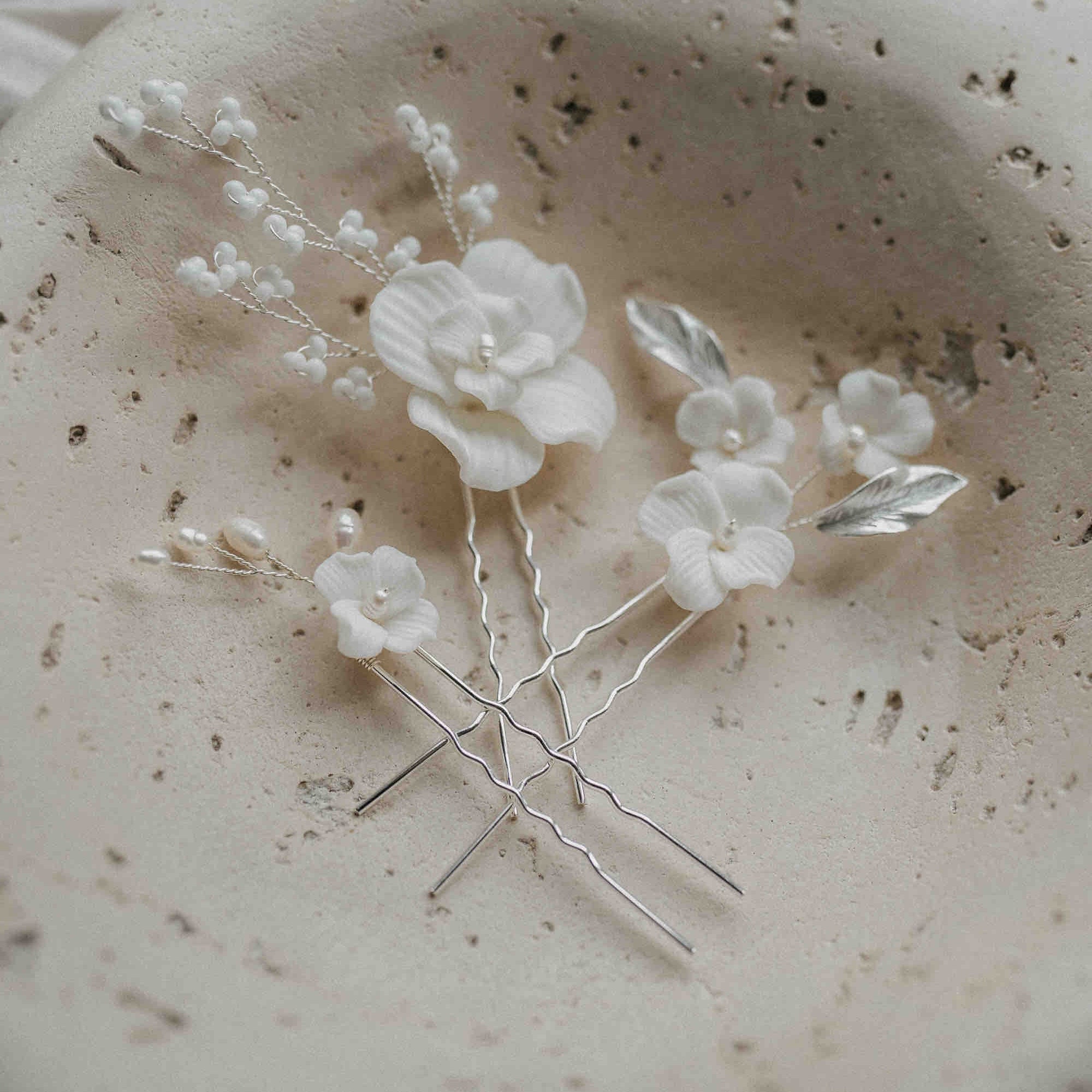 Drei Haarnadeln mit Keramik Blüten und Perlen (3 Stück) von Fräulein Maya mit weißen Blüten, Perlen und silbernen Blättern liegen in einer Schale - ideal für eine romantische Brautfrisur.