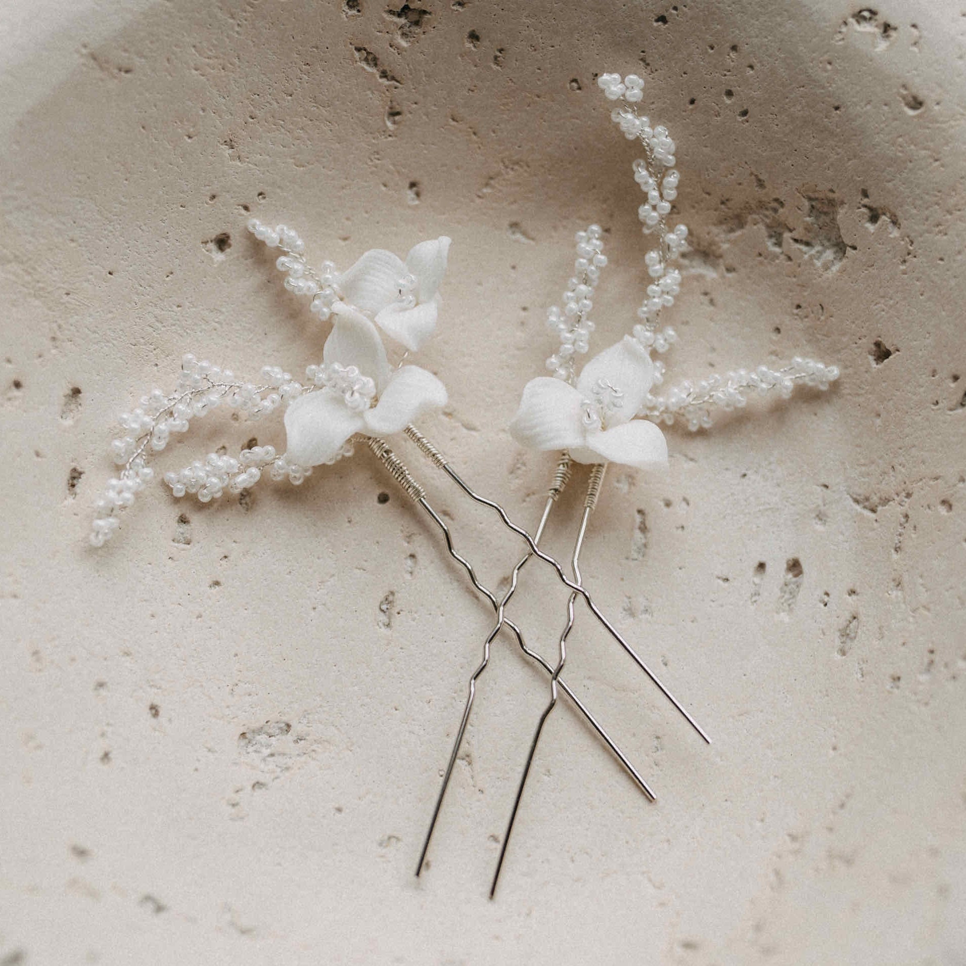 Zwei Keramik Haarnadeln mit Blumen und feinen Perlen von Fräulein Maya (2 Stück) liegen auf einer strukturierten, hellbeigen Steinoberfläche mit natürlichen Löchern und Unregelmäßigkeiten.