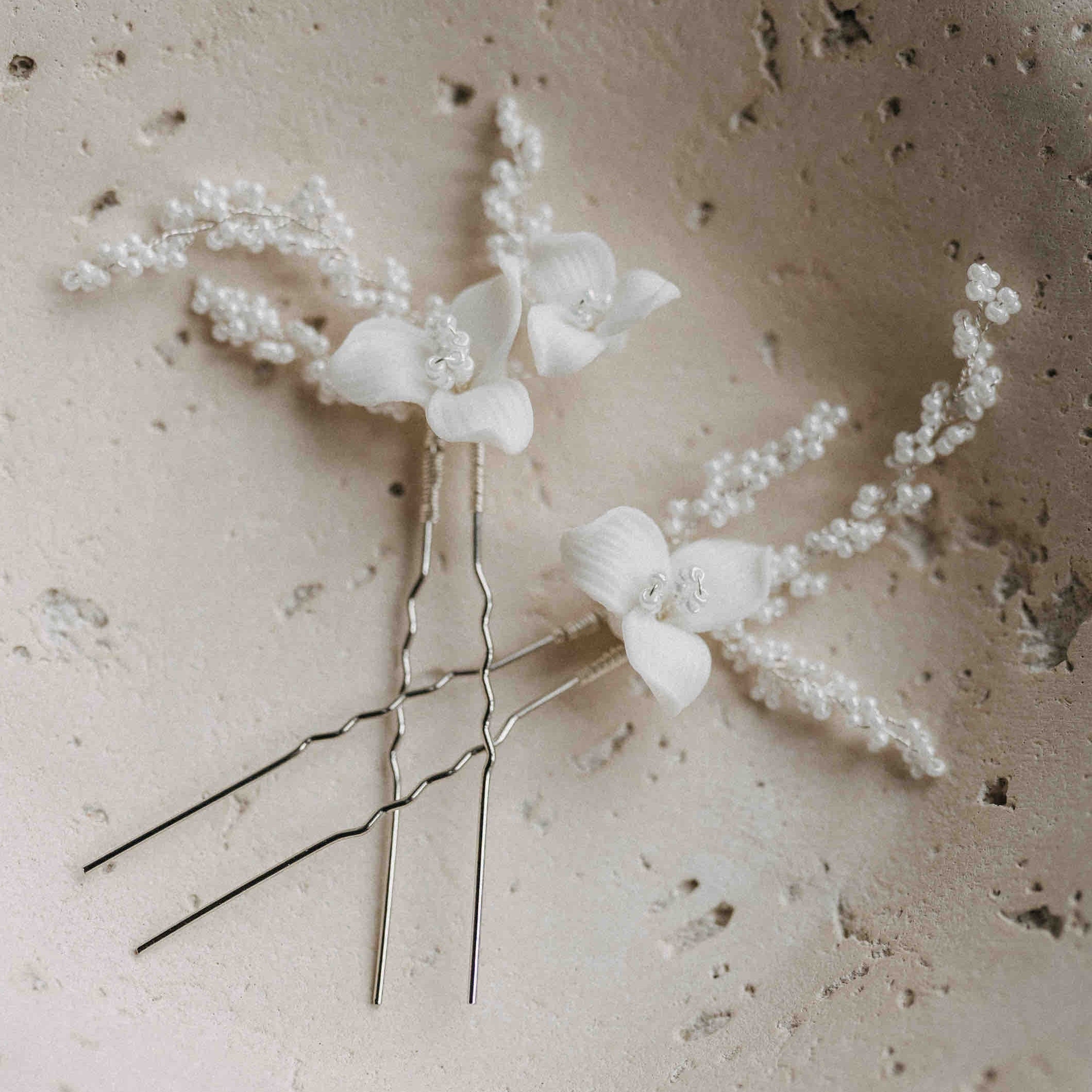 Zwei "Keramik Haarnadeln mit Blumen und feinen Perlen" (2 Stück) von Fräulein Maya, verziert mit keramischen Blüten und kleinen Perlen, liegen parallel auf einer strukturierten, hellen Steinoberfläche.
