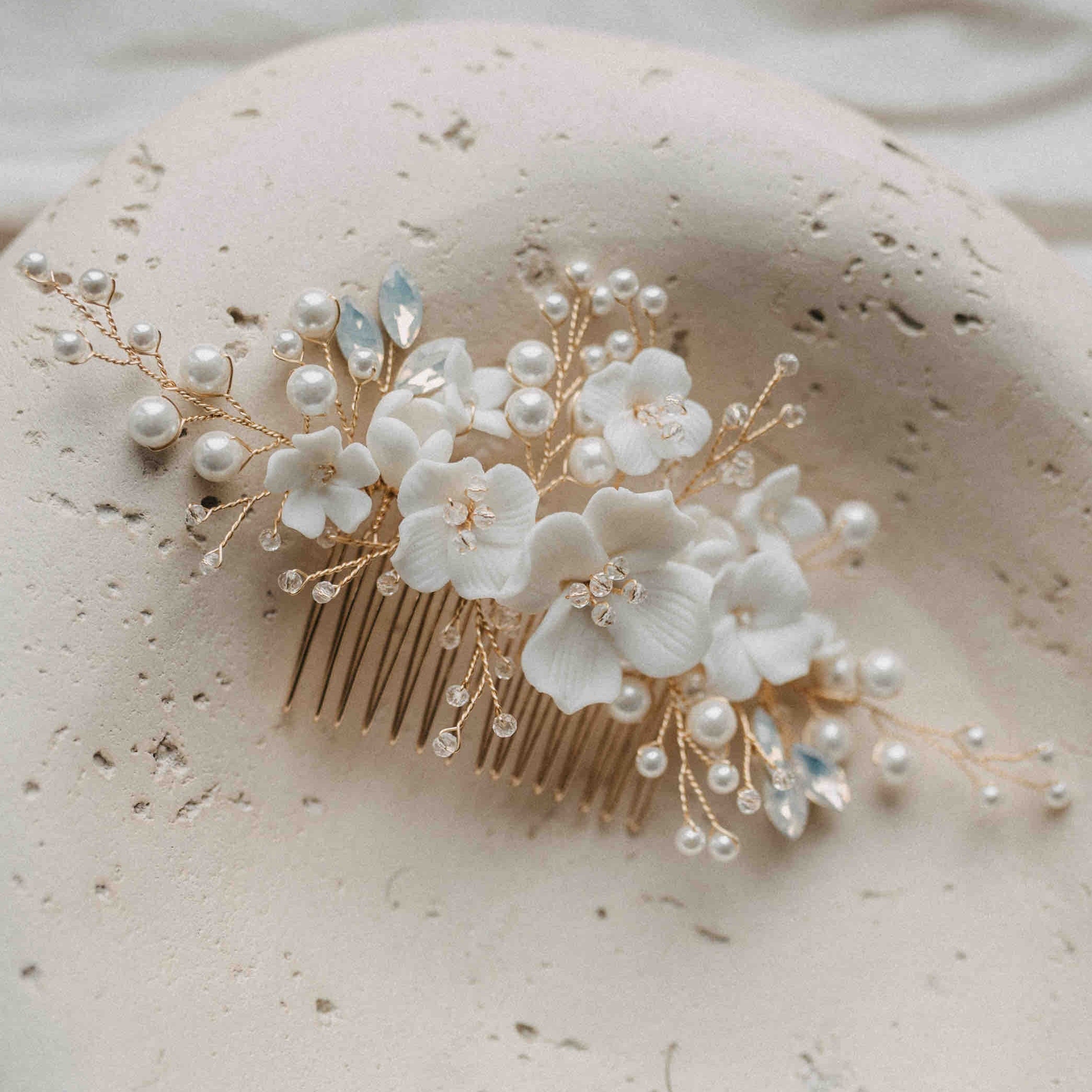 Der Haarkamm mit Keramik Blumen und Kristallen von Fräulein Maya besticht durch Keramikblumen, weiße Blüten, Glasperlen und kleine Kristalle-ideal als Braut-Accessoire auf hellem Stein im sanften Licht arrangiert.