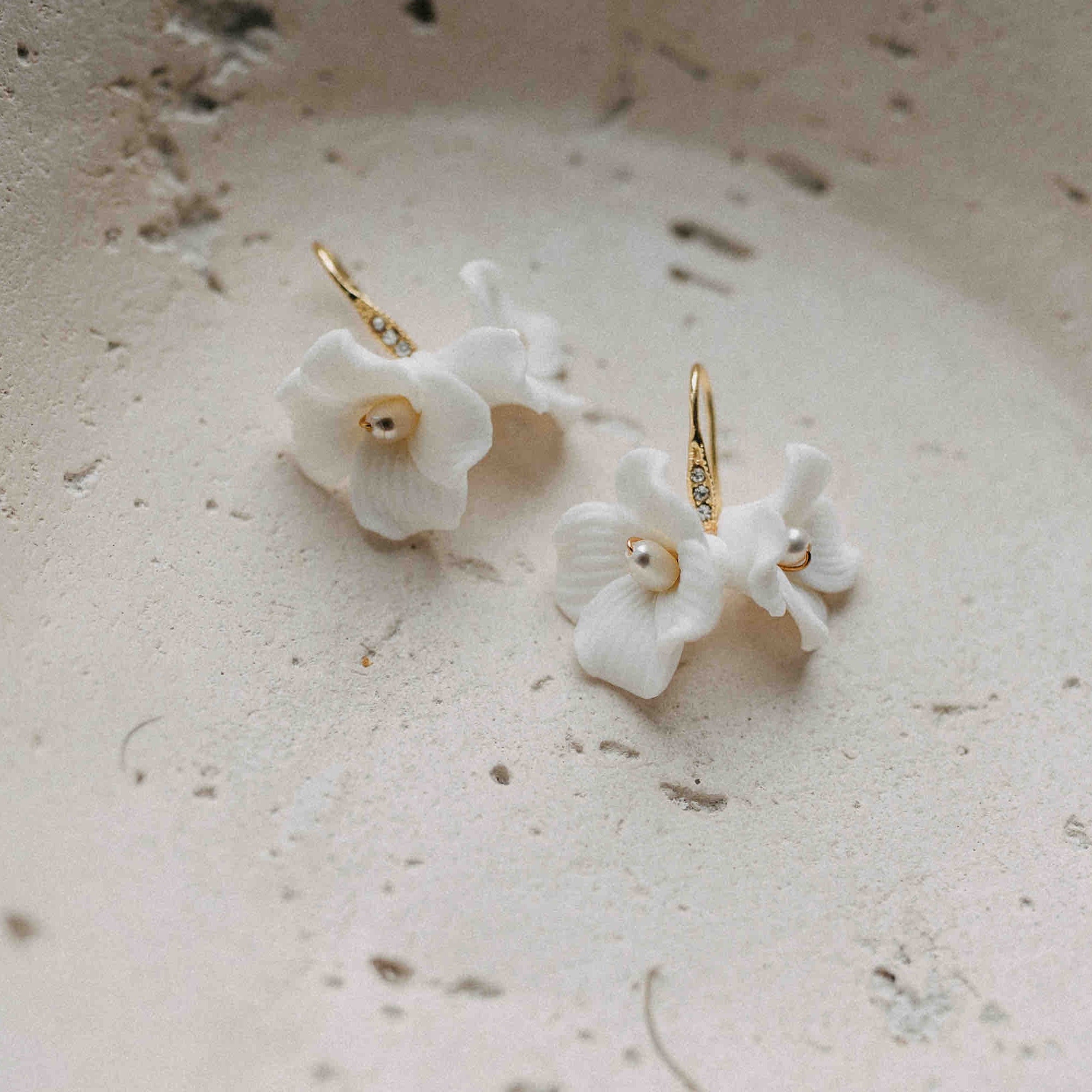 Ein Paar Braut Ohrringe mit Keramik Blüten von Fräulein Maya ruht auf einer hellbeigen Oberfläche. Jeder Ohrring hat eine weiße Keramikblüte mit Perlen in der Mitte und goldenen Haken - ideal als Boho-Brautaccessoire für eine elegante Frisur.