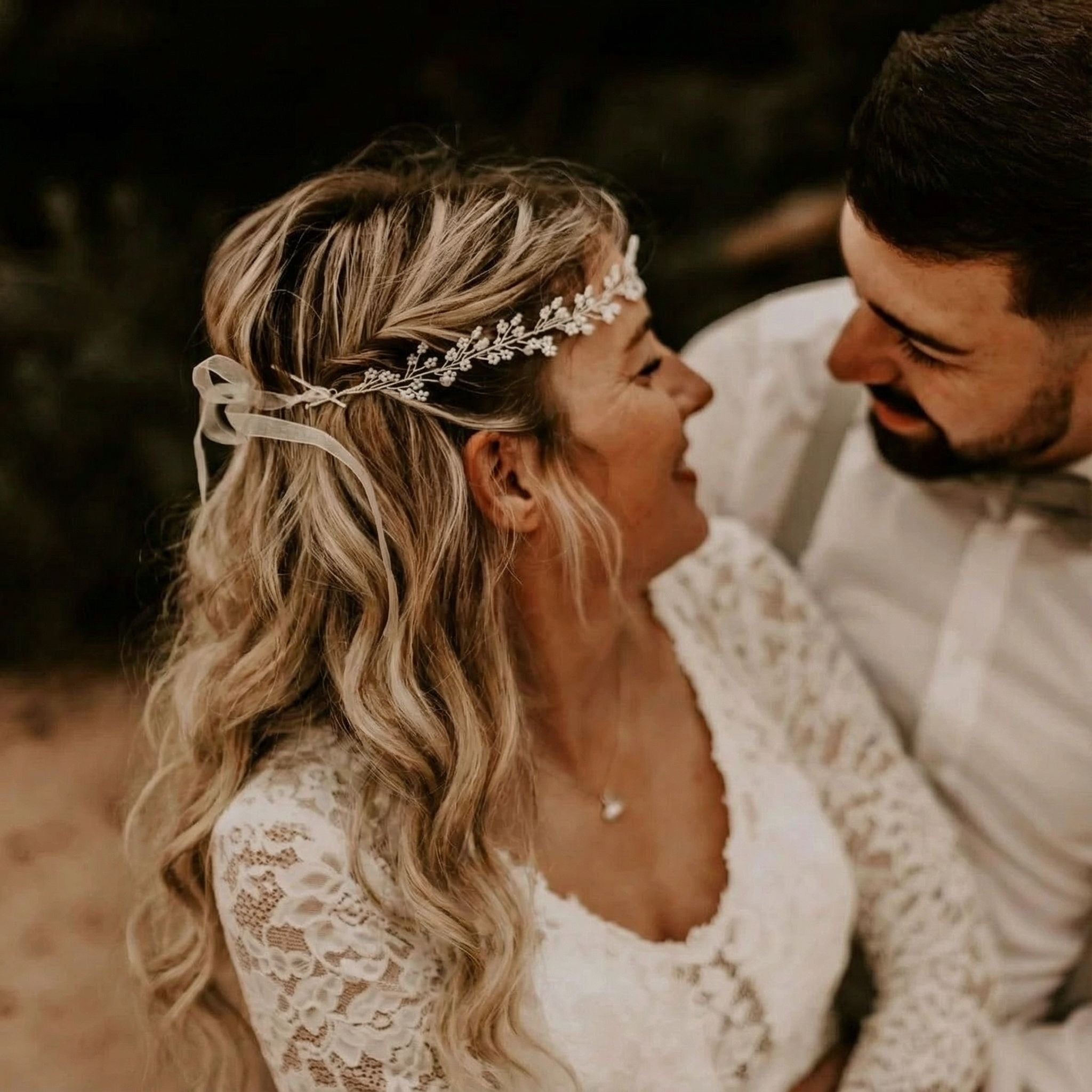 Eine Braut in einem Spitzenkleid und einem schmuckvollen Kopfschmuck lächelt einen Mann neben ihr an. Beide haben helle Haut und gewelltes Haar; er trägt ein weißes Hemd und eine Fliege. Das Paar verbringt einen freudigen, intimen Moment im Freien bei weichem, natürlichem Licht.