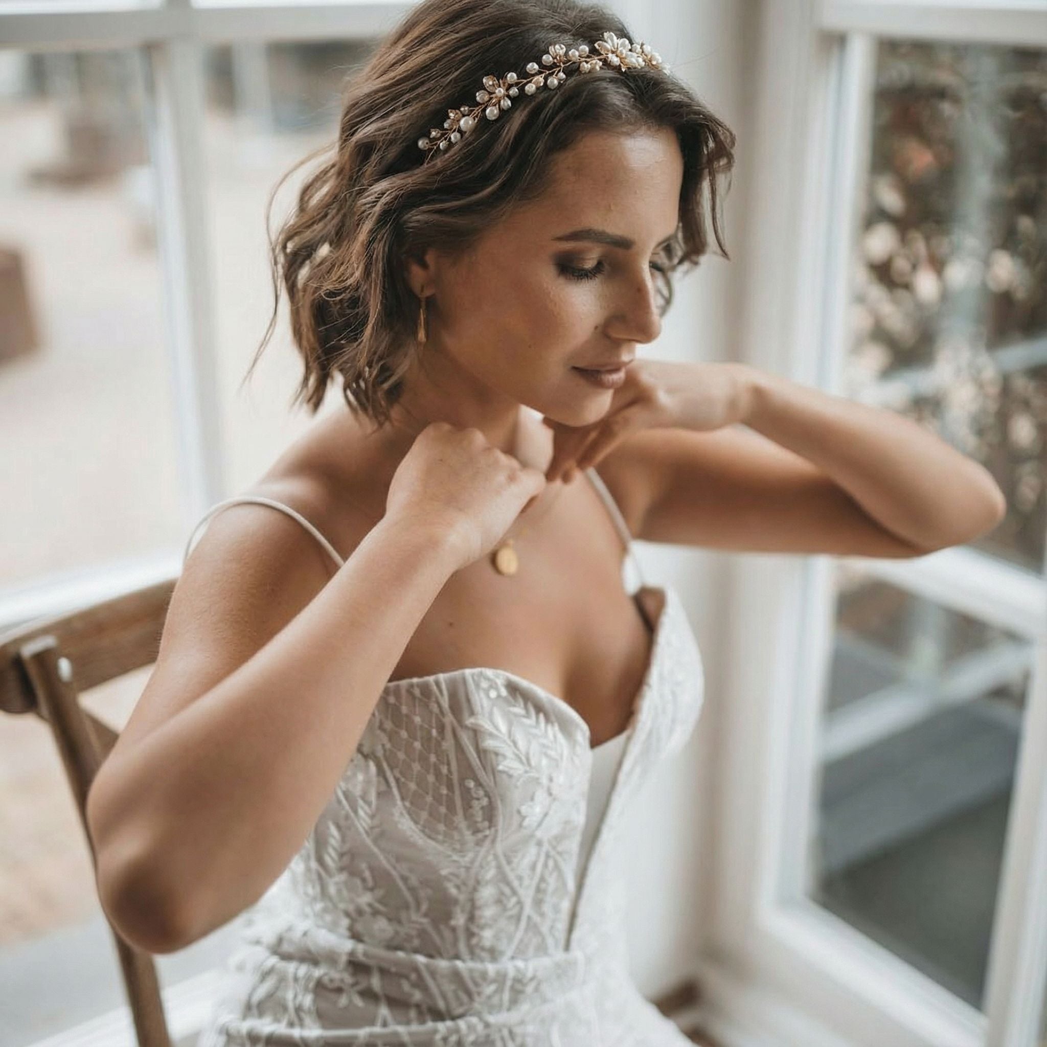 Eine Frau in einem weißen, bestickten Brautkleid sitzt an einem Fenster und rückt eine goldene Halskette zurecht. Sie hat kurzes braunes Haar, das in Wellen frisiert ist, und trägt ein zartes Blumenstirnband. Weiches natürliches Licht beleuchtet ihren heiteren Ausdruck und ihre elegante Kleidung.