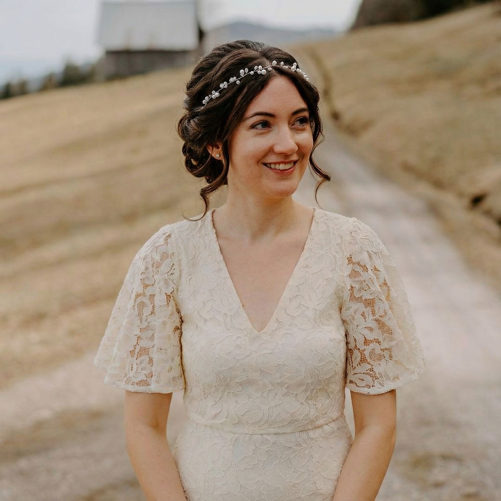 Eine Frau in einem cremefarbenen Spitzenkleid mit Flatterärmeln steht auf einem Feldweg im Freien. Sie hat braunes Haar, das mit einem juwelenbesetzten Stirnband hochgesteckt ist, lächelt und schaut leicht zur Seite. Im Hintergrund sind ein grasbewachsener Hügel und eine rustikale Scheune zu sehen.