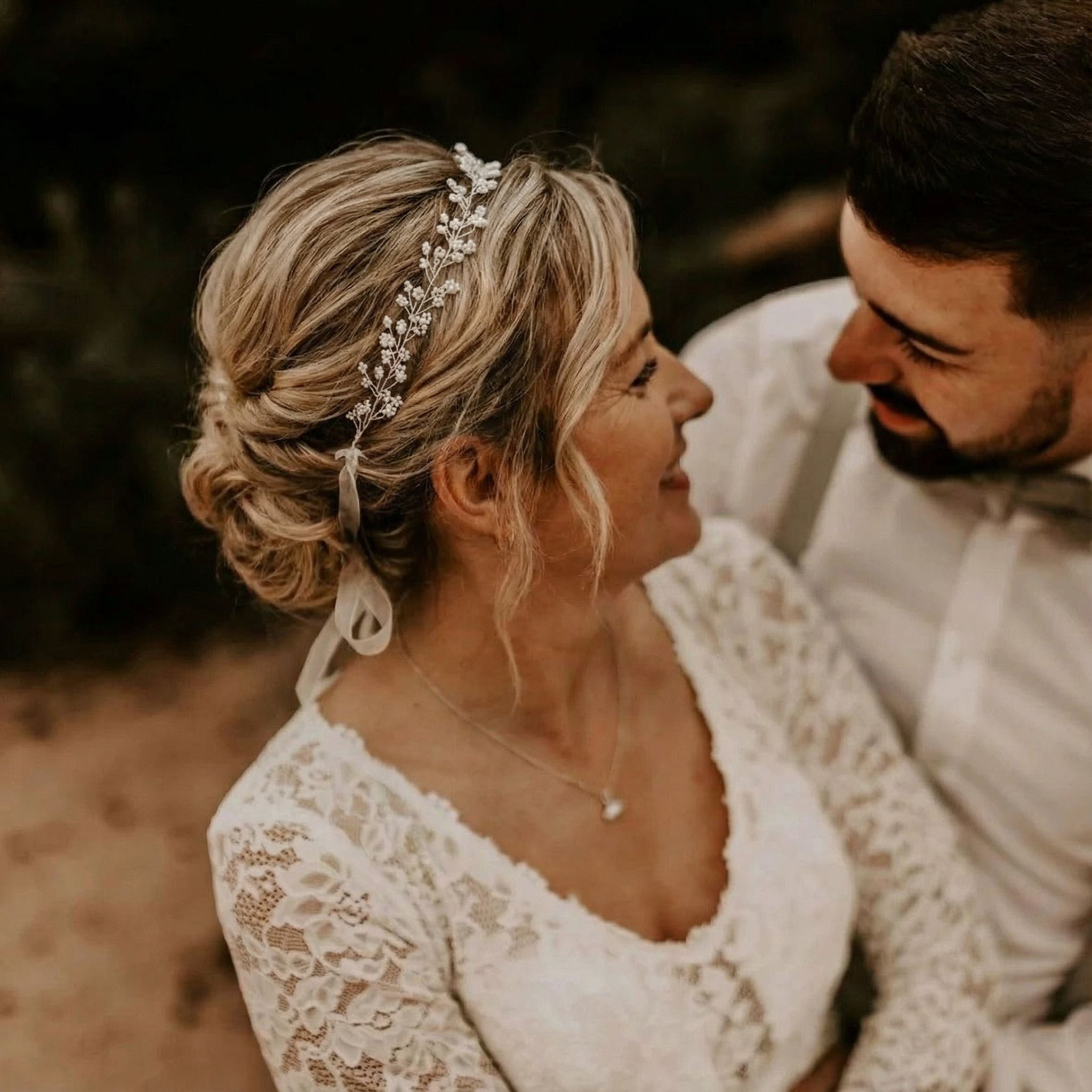 Eine Frau in einem weißen Spitzenhochzeitskleid und einem schmuckvollen Stirnband lächelt einen bärtigen Mann in einem hellen Hemd und einer Fliege an. Sie stehen sich nahe gegenüber und teilen einen freudigen, intimen Moment. Der Hintergrund ist unscharf, um ihre Verbindung hervorzuheben.