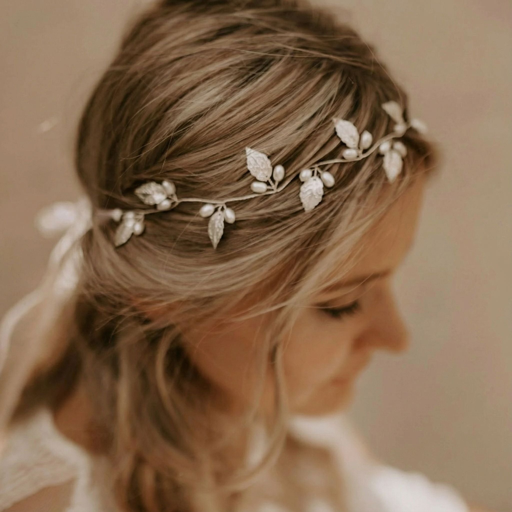 Eine Frau mit hellbraunem, gewelltem Haar ist in Weichzeichner dargestellt und blickt nach unten. Sie trägt eine zarte silberne Kopfbedeckung, die mit Blättern und Perlen verziert ist. Der Hintergrund ist neutral und schafft eine verträumte, ätherische Atmosphäre.