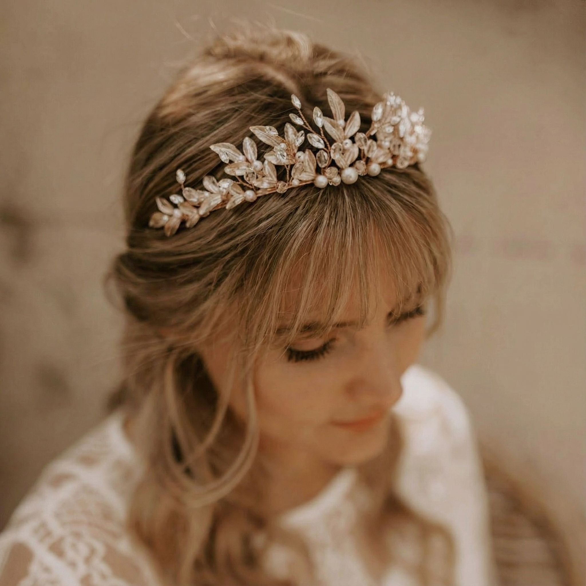 Eine Frau mit gewelltem blondem Haar, das halb hochgesteckt ist, trägt ein zartes, funkelndes Diadem, das mit Perlen und kristallähnlichen Steinen besetzt ist. Sie hat einen weichen Pony, lange Wimpern und ist in ein weißes Spitzenkleid gekleidet, das in einer warmen, eleganten Umgebung sanft beleuchtet wird.