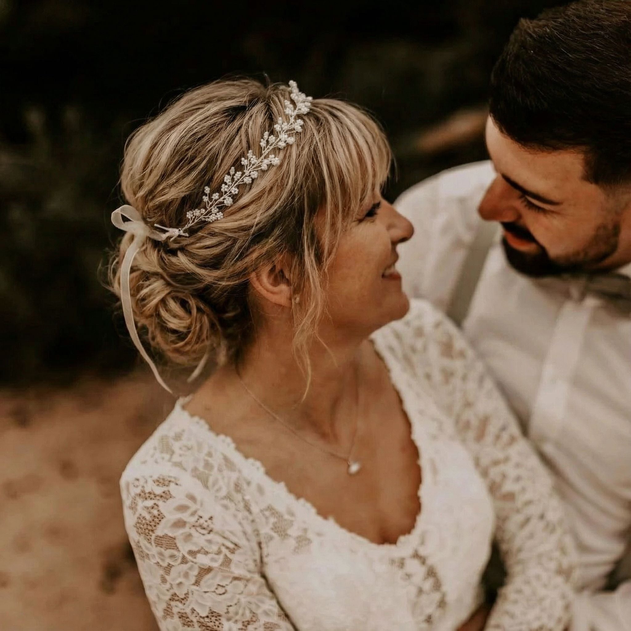 Eine lächelnde Braut mit juwelenbesetztem Haarschmuck und Spitzenkleid blickt liebevoll auf einen Bräutigam mit Bart und Fliege. Sie stehen eng beieinander und halten sich gegenseitig in einer warmen, sanft beleuchteten Umgebung im Freien.