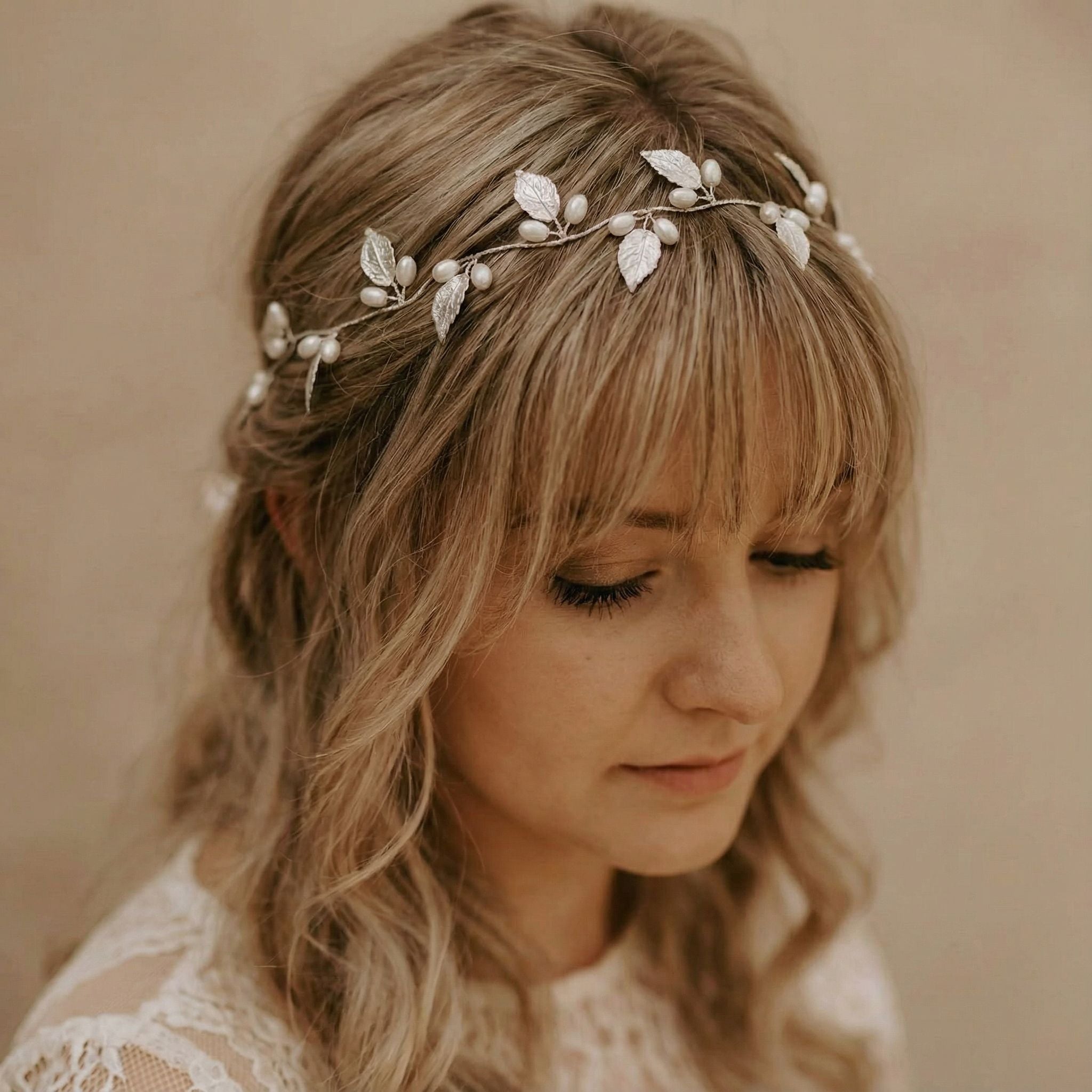 Eine Frau mit gewelltem blondem Haar und Pony trägt eine zarte Kopfbedeckung, die mit silbernen Blättern und Perlen verziert ist. Sie ist mit einem weißen Spitzenkleid bekleidet und blickt vor einem neutralen beigen Hintergrund mit heiterem Blick nach unten.
