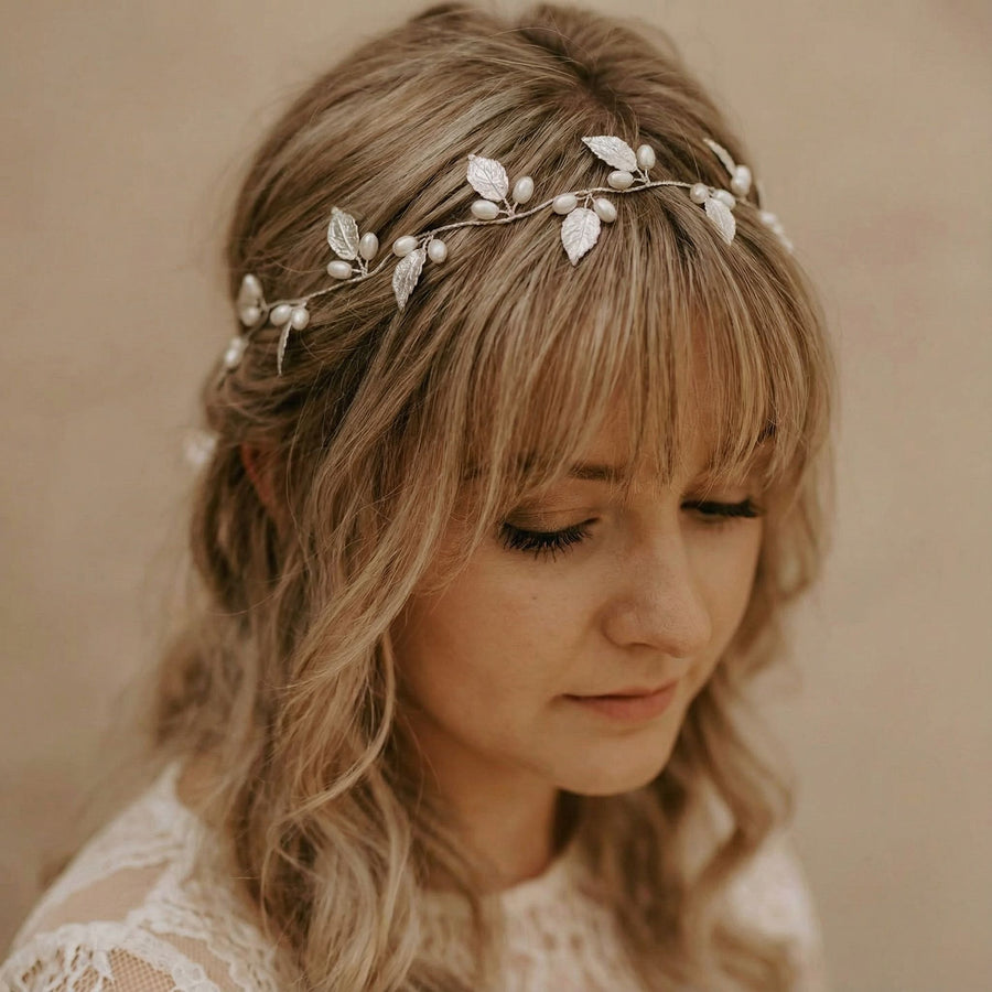 Eine Frau mit gewelltem blondem Haar und Pony trägt eine zarte Kopfbedeckung, die mit silbernen Blättern und Perlen verziert ist. Sie ist mit einem weißen Spitzenkleid bekleidet und blickt vor einem neutralen beigen Hintergrund mit heiterem Blick nach unten.