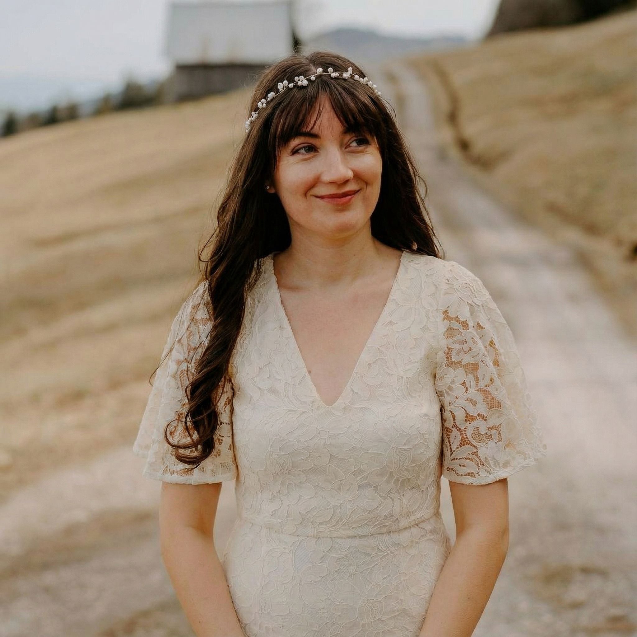 Eine Frau mit langen braunen Haaren, die ein weißes Spitzenkleid und ein zartes Perlenstirnband trägt, steht auf einem Feldweg. Sie lächelt sanft, im Hintergrund sind eine unscharfe rustikale Scheune und grasbewachsene Felder an einem bewölkten Tag zu sehen.