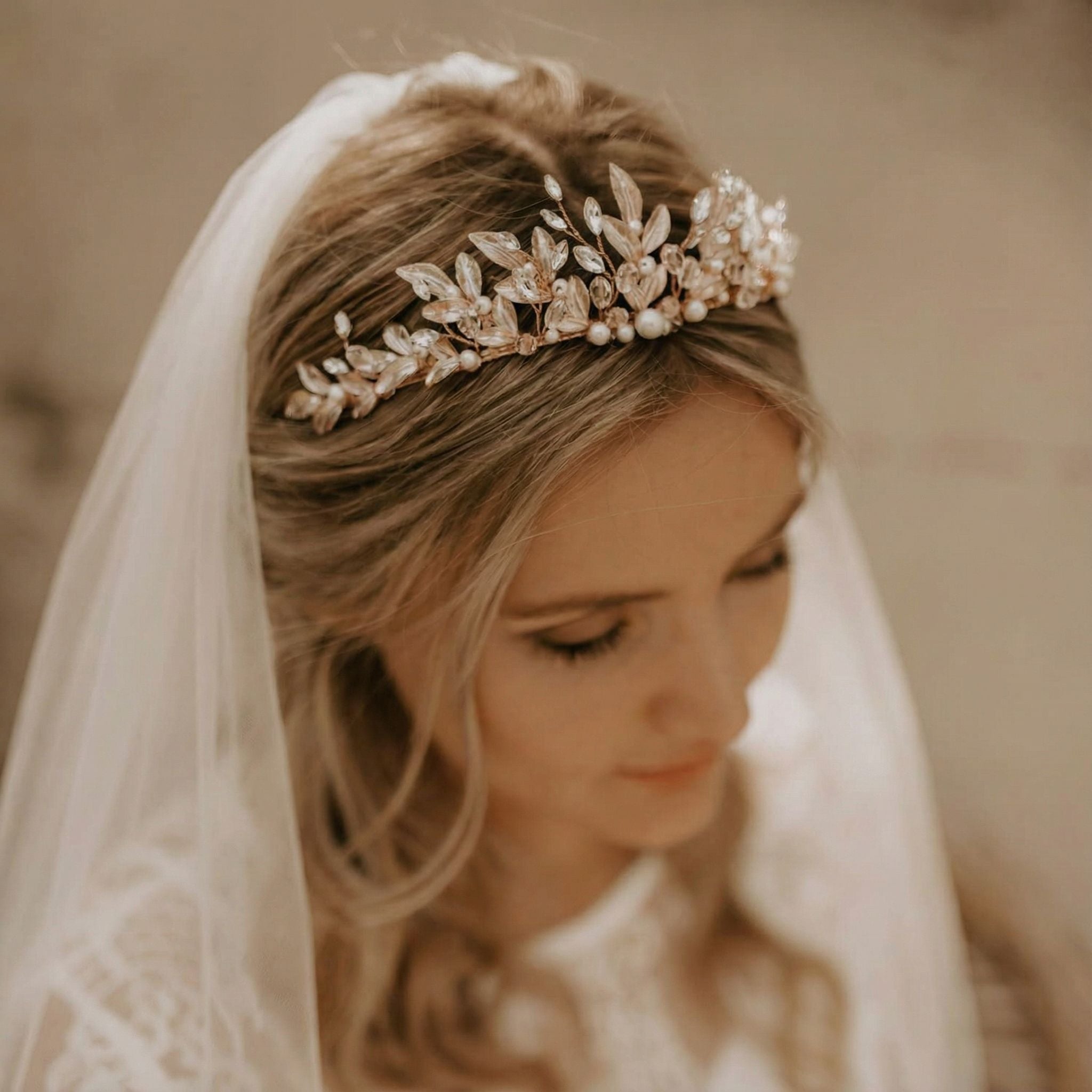 Eine Braut mit gewelltem blondem Haar trägt ein zartes, perlenbesetztes Blumen-Diadem und einen weißen Schleier. Sie blickt sanft nach unten, ihr kunstvolles Spitzenkleid ist teilweise sichtbar, und die warme, sanfte Beleuchtung schafft eine romantische und elegante Atmosphäre.