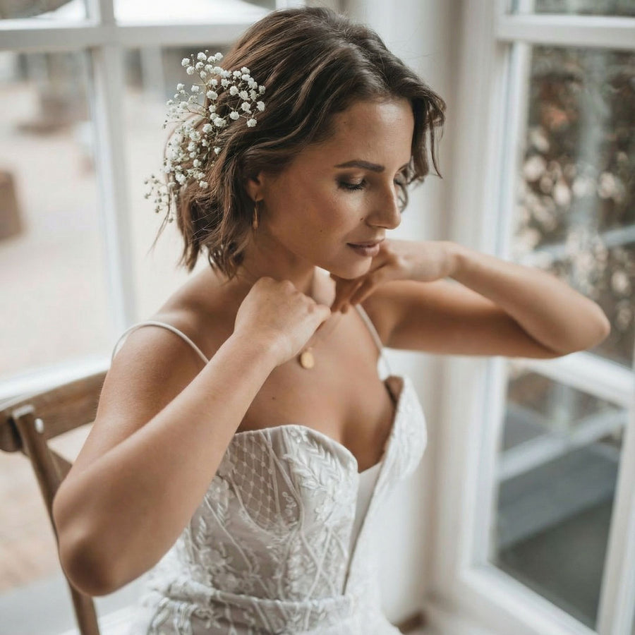 Eine Frau in einem weißen, blumenbestickten Brautkleid sitzt auf einem Holzstuhl an einem großen Fenster. Sie hat schulterlanges braunes Haar, in das zarte weiße Blumen gesteckt sind, und berührt sanft ihre Halskette, während sie mit heiterer Miene nach unten schaut.