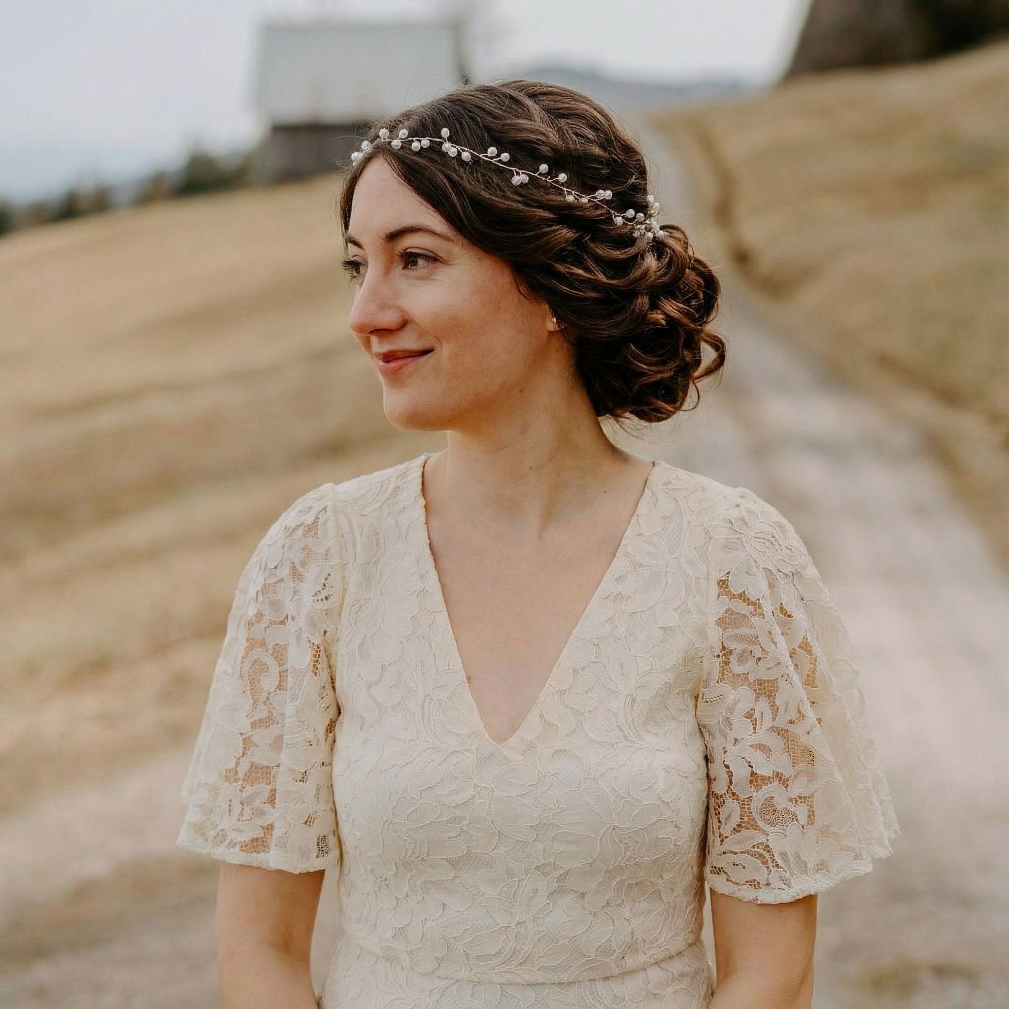 Eine Frau in einem cremefarbenen Spitzenkleid mit Flatterärmeln steht im Freien auf einem Weg und schaut nach links. Ihr braunes Haar ist zu einer eleganten Hochsteckfrisur mit einer Perlenkette frisiert. Der Hintergrund ist ein unscharfes, grasbewachsenes Feld mit einer Scheune in der Ferne.