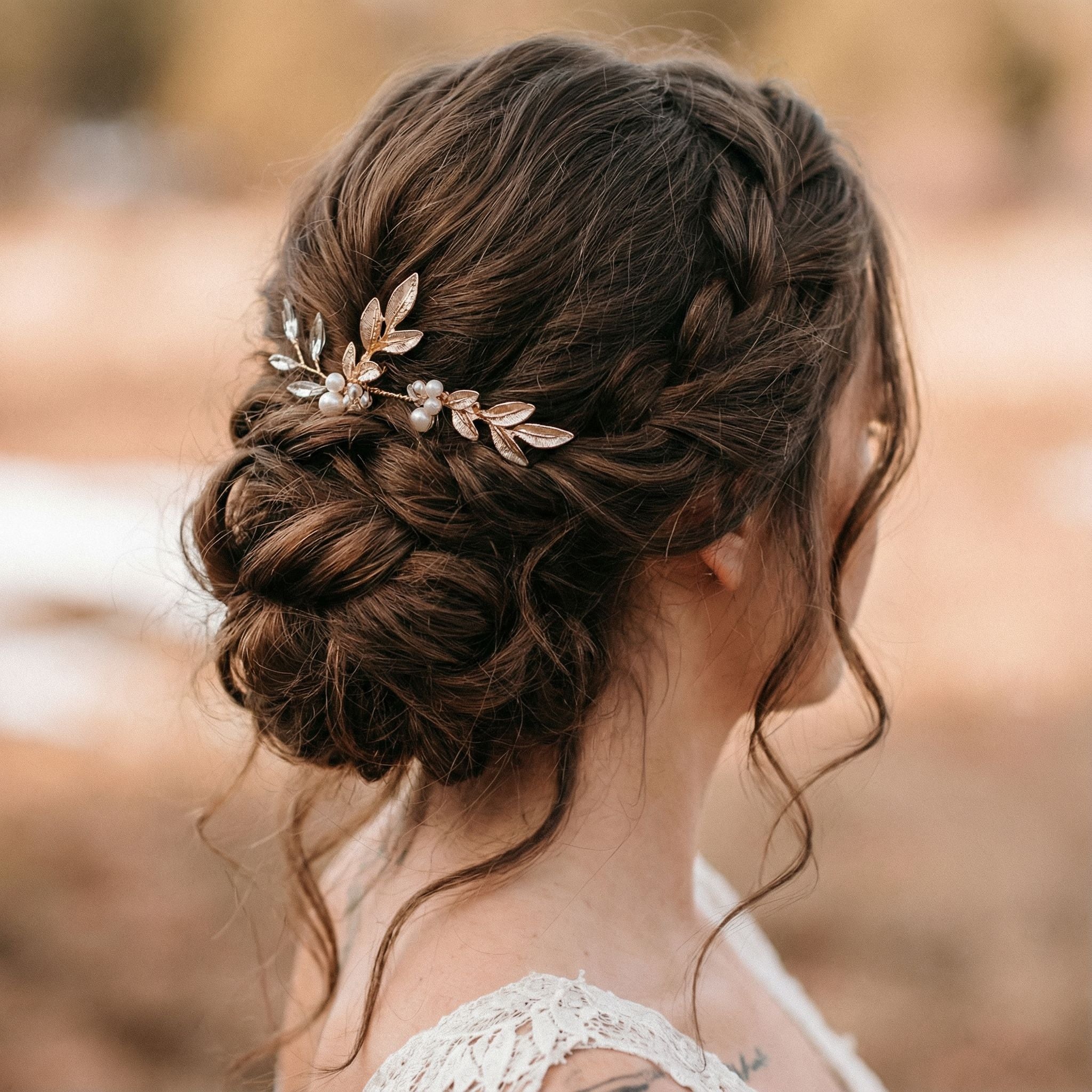 Eine Frau mit langen braunen Haaren, die zu einer niedrigen, locker geflochtenen Hochsteckfrisur frisiert sind, wobei weiche Locken ihr Gesicht umrahmen. Ihr Haar ist mit einem Haarschmuck aus Blattgold und Perlen geschmückt. Sie trägt ein Spitzenkleid und steht im Freien vor einem unscharfen, warm getönten Hintergrund.