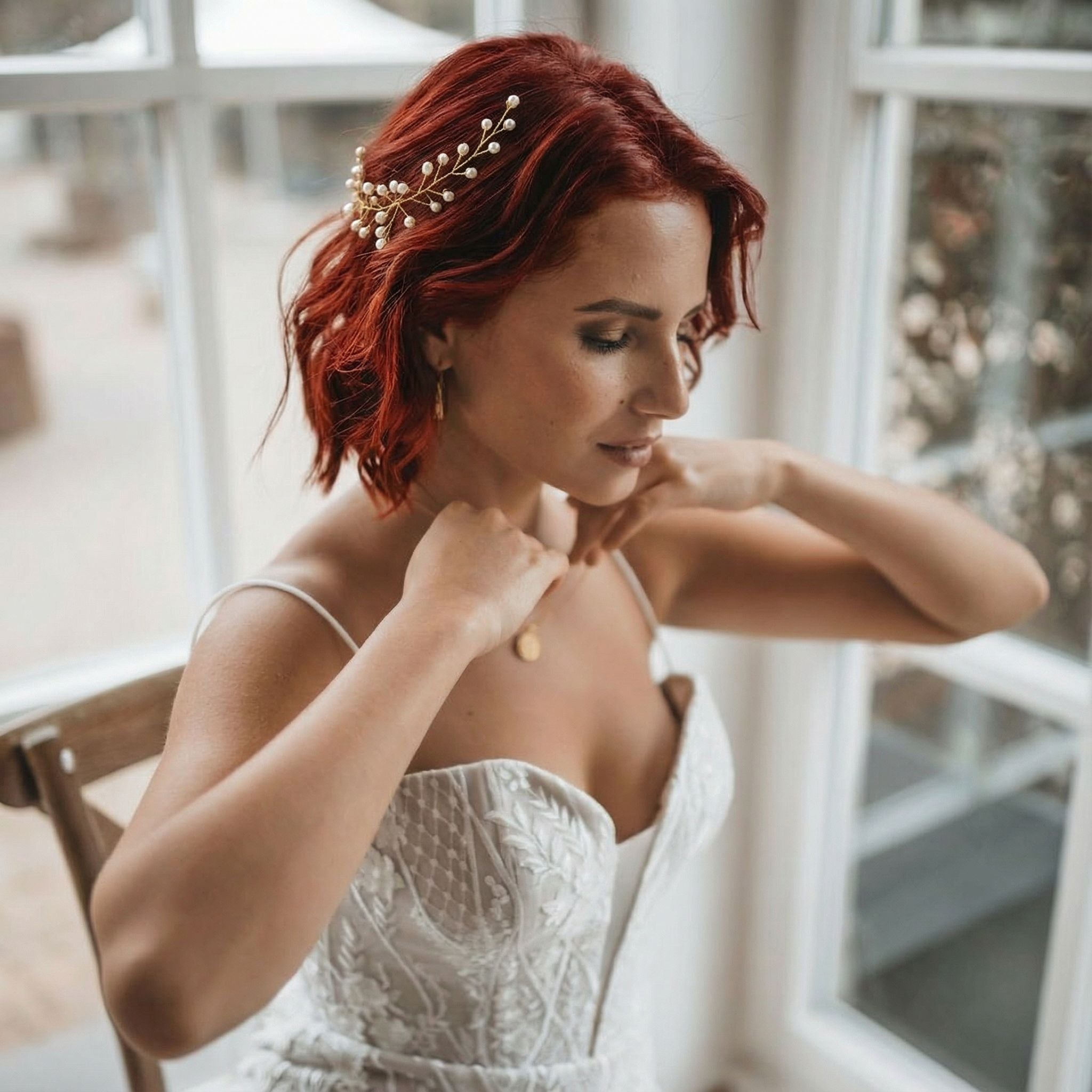 Eine Frau mit rotem Haar, das in lockeren Wellen gestylt und mit einem goldenen Haarteil geschmückt ist, rückt eine zarte Halskette zurecht. Sie trägt ein weißes, trägerloses, strukturiertes Hochzeitskleid und sitzt an einem sonnenbeschienenen Fenster, gelassen und nachdenklich wirkend.