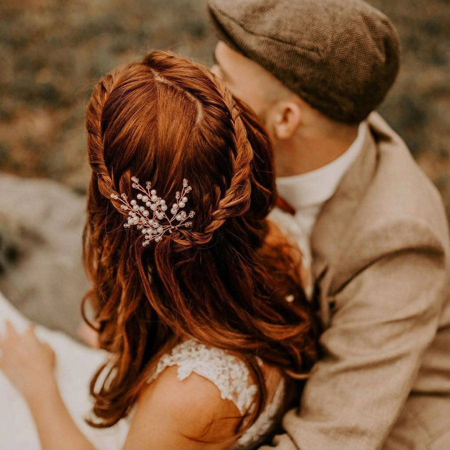Eine Frau mit gewelltem, kastanienbraunem Haar, das zu Zöpfen geflochten und mit einem filigranen Perlenhaarschmuck verziert ist, sitzt dicht neben einem Mann in einem braunen Tweedanzug und einer flachen Mütze. Beide sind einander zugewandt und halten sich im Freien in warmem, weichem Licht.